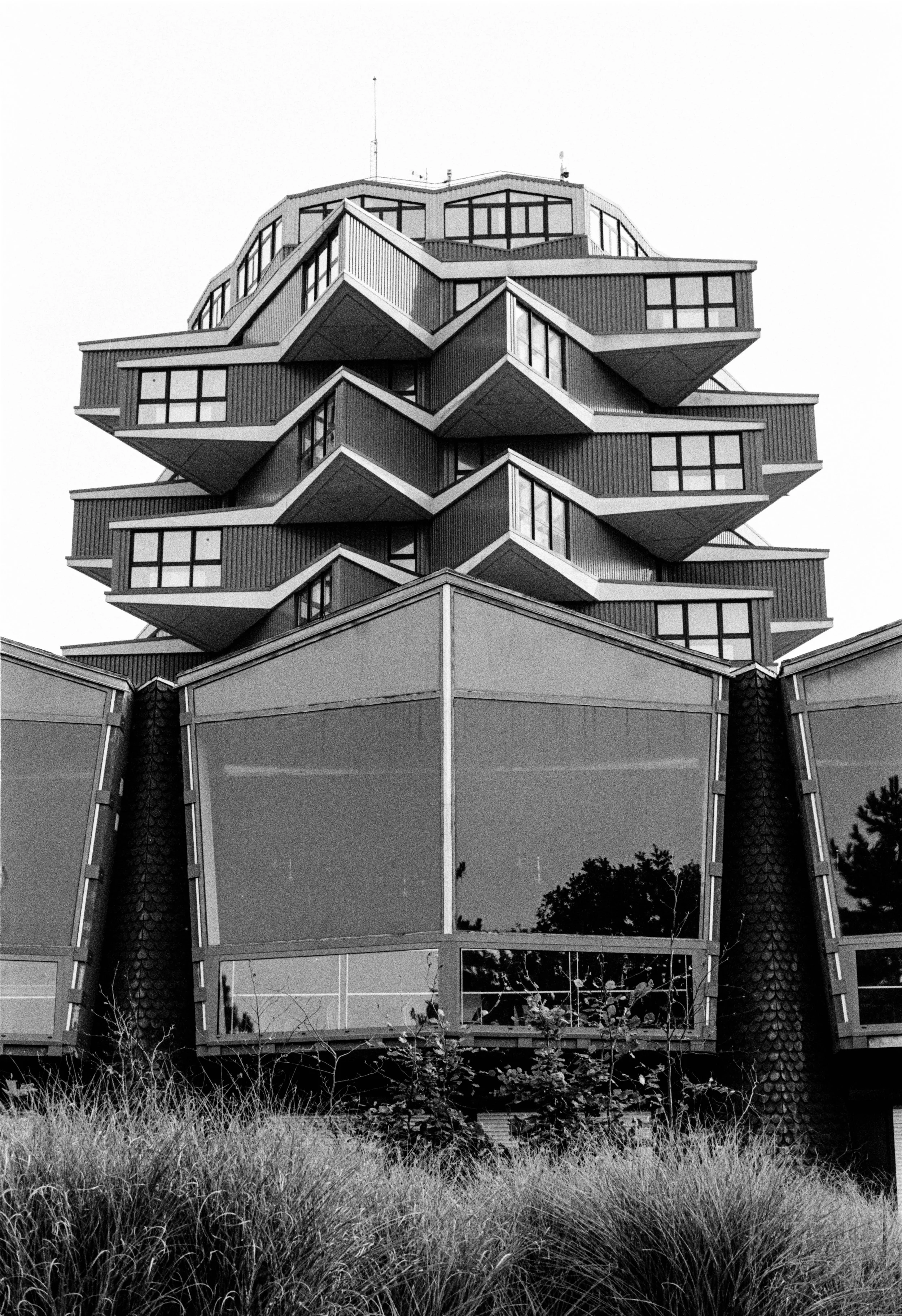 Un bâtiment moderne avec une architecture inhabituelle, avec des niveaux en saillie disposés de manière décalée et des fenêtres rectangulaires. La photo est en noir et blanc, et des arbustes sont visibles en premier plan.