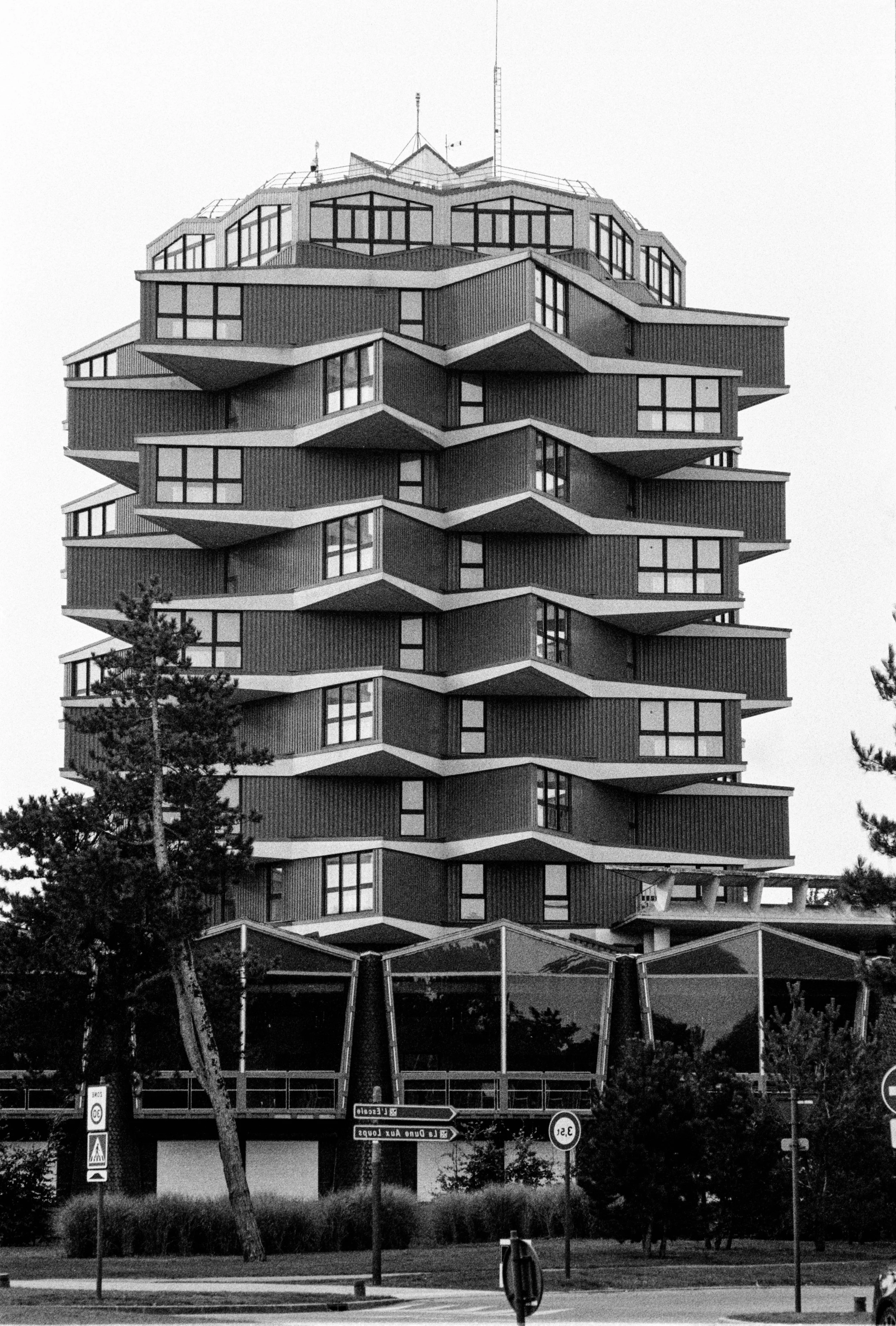 Un bâtiment moderne à plusieurs étages avec une architecture unique en forme de vagues ou d'escaliers, composé principalement de fenêtres rectangulaires et d'une façade en panneaux. Il est entouré d'arbres et de panneaux de signalisation routière.