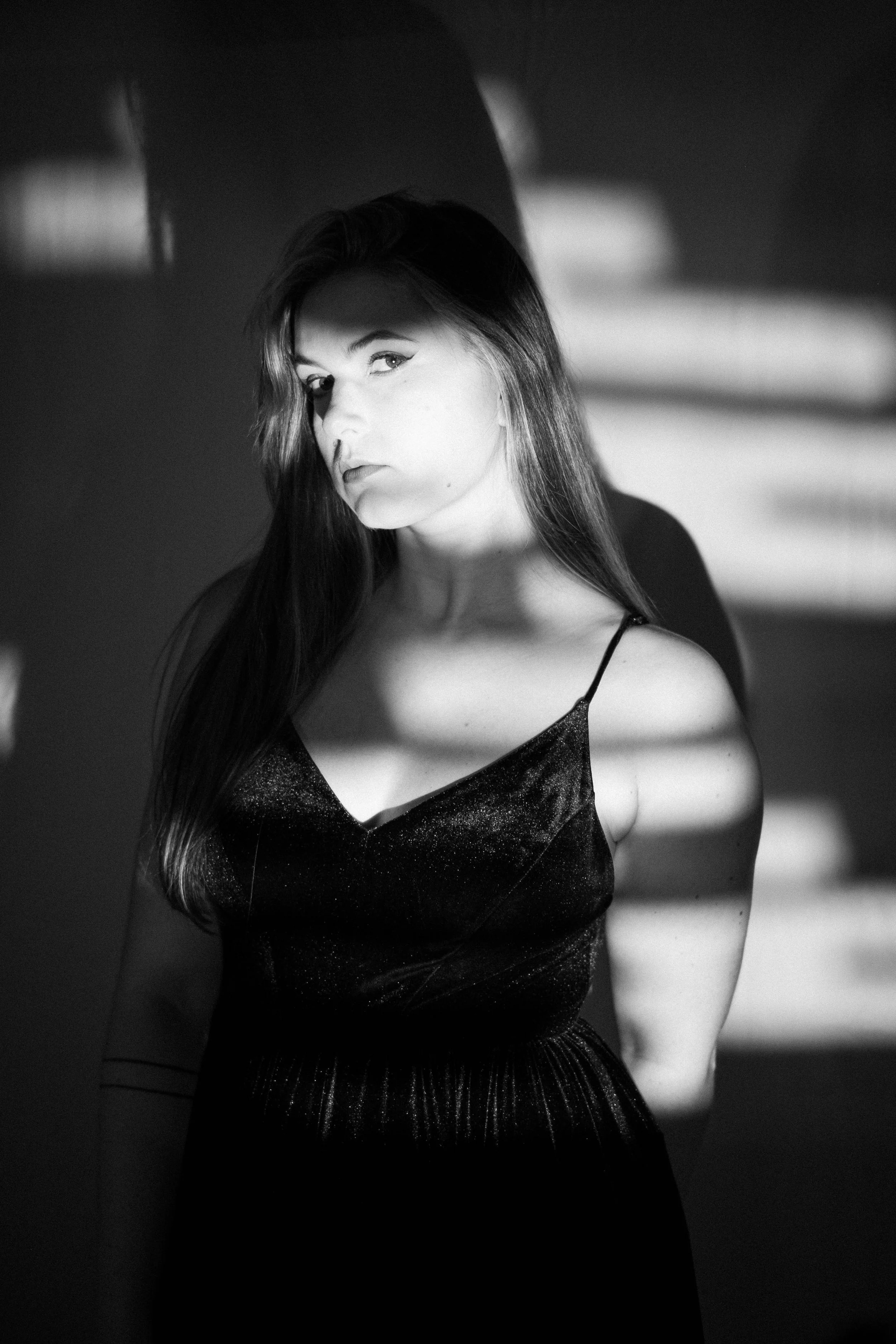 Une femme en dress noir, en noir et blanc, regardant vers l'objectif dans un jeu d'ombres et de lumières.