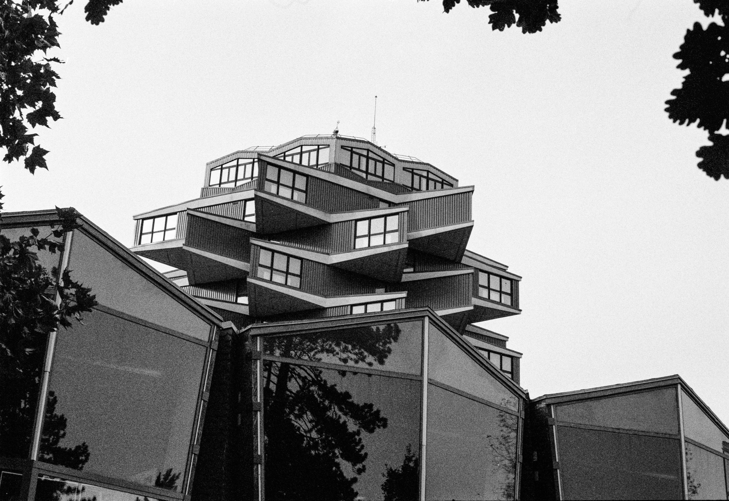 Photo en noir et blanc d'un bâtiment moderne avec plusieurs étages de structures en saillie et grandes fenêtres, entouré d'arbres.