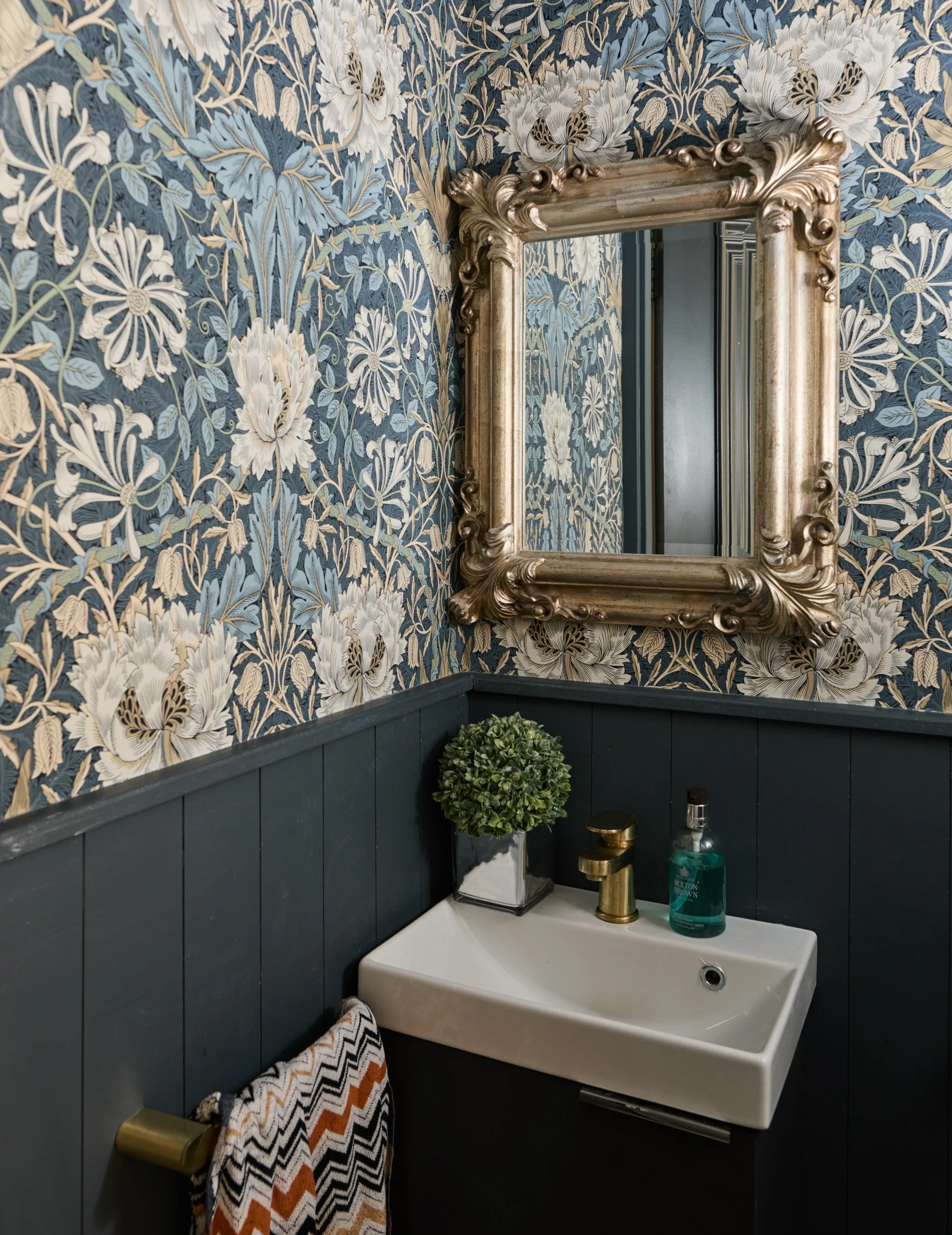 A bathroom corner with floral patterned wallpaper, a gold-framed mirror, a corner sink with a gold faucet, a small potted plant, a blue-green liquid soap dispenser, and a towel with a black, white, and orange zigzag pattern hanging on a gold towel ri