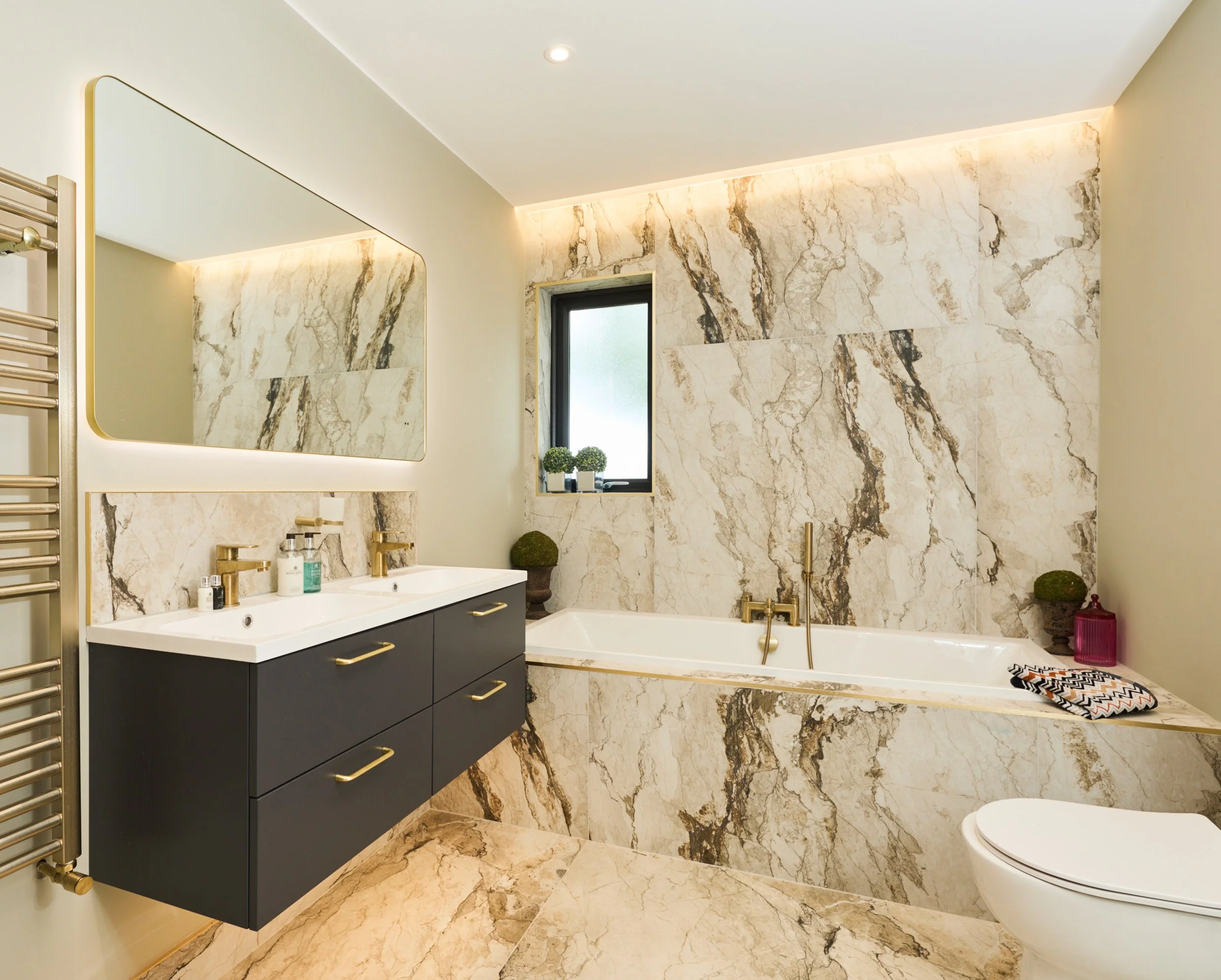 Modern bathroom with a marble accent wall and floor, a white bathtub with gold fixtures, a double vanity with gold handles, a large mirror, a window with small potted plants, and a wall-mounted towel warmer.