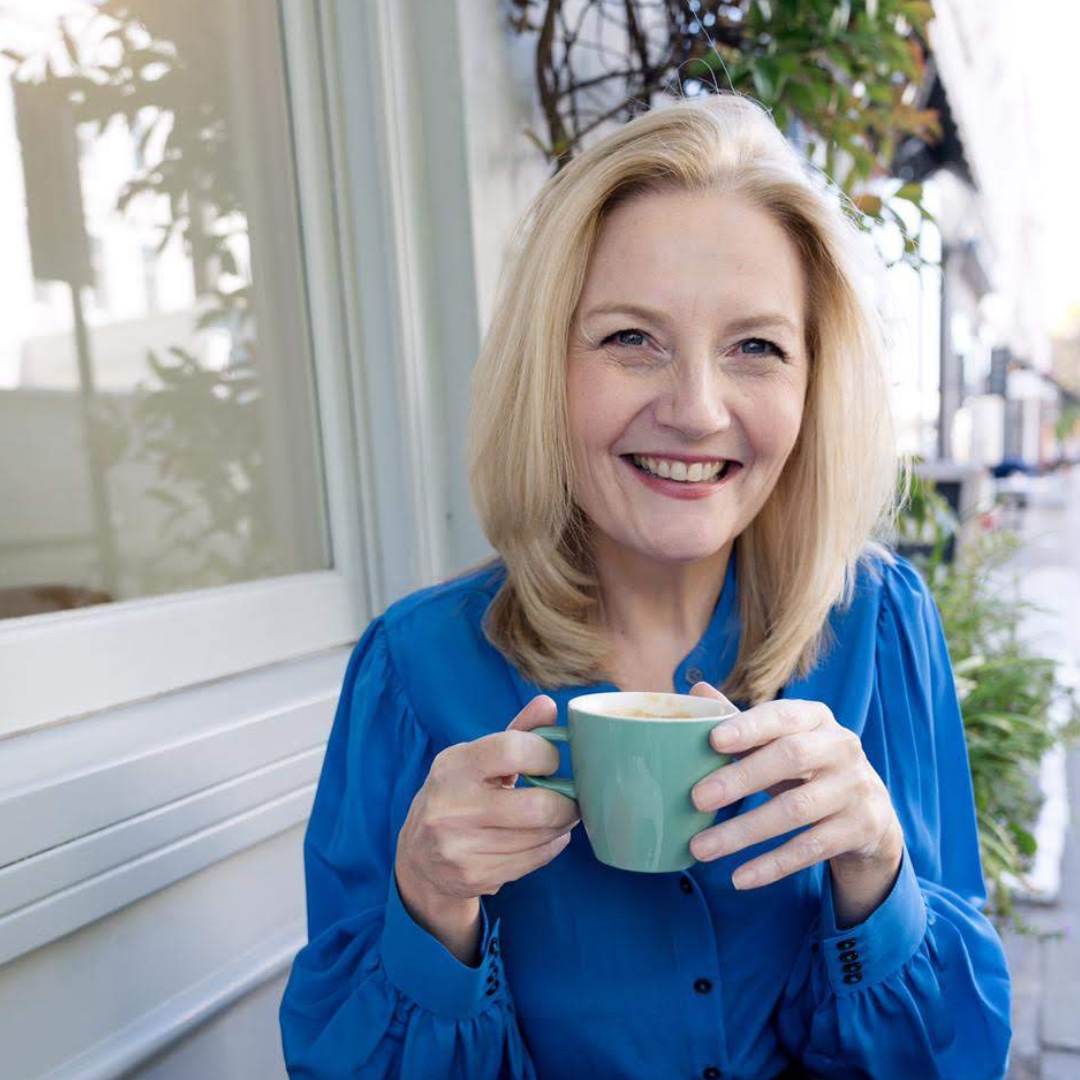 A woman with blonde hair smiling and holding a blue mug outdoors, sitting near a window with plants in the background.