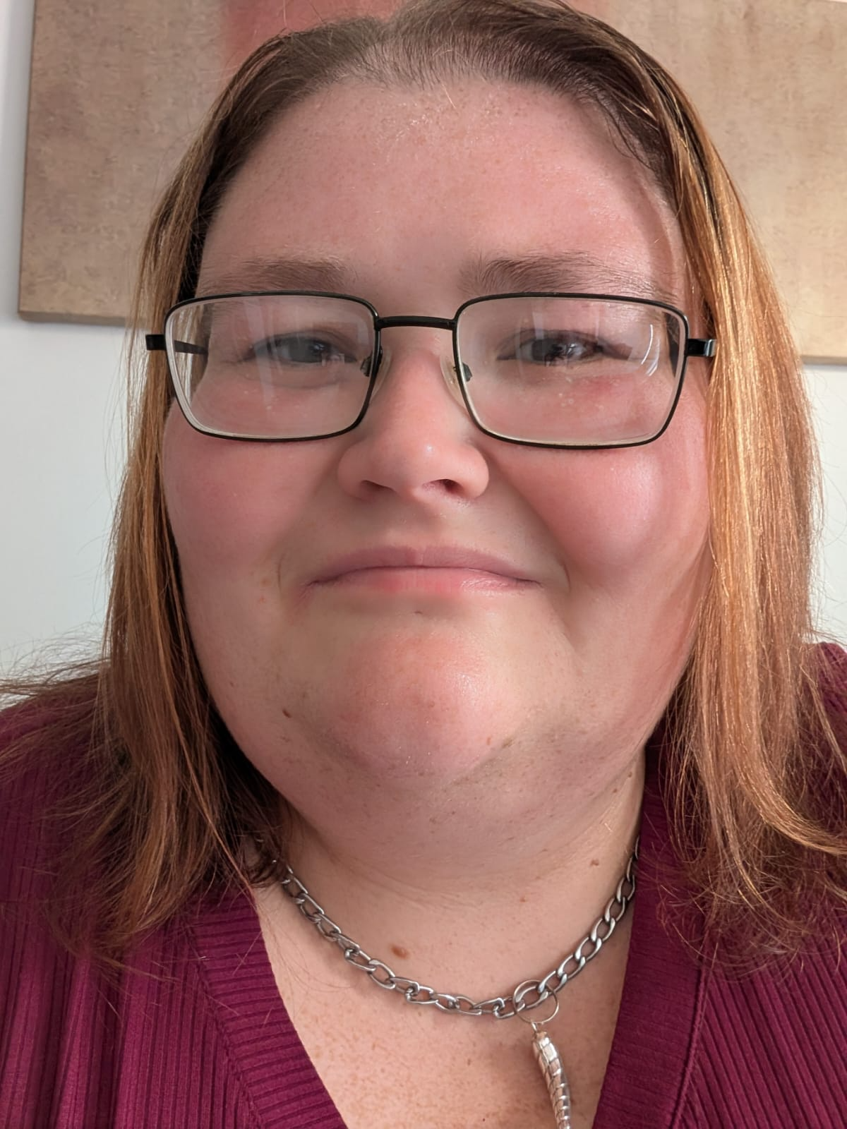 Close-up of a smiling woman with red hair, glasses, and a silver chain necklace, wearing a maroon top.