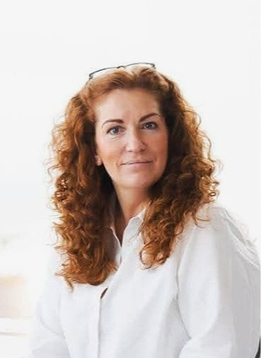 A woman with curly red hair wearing a white shirt, looking at the camera with a slight smile.