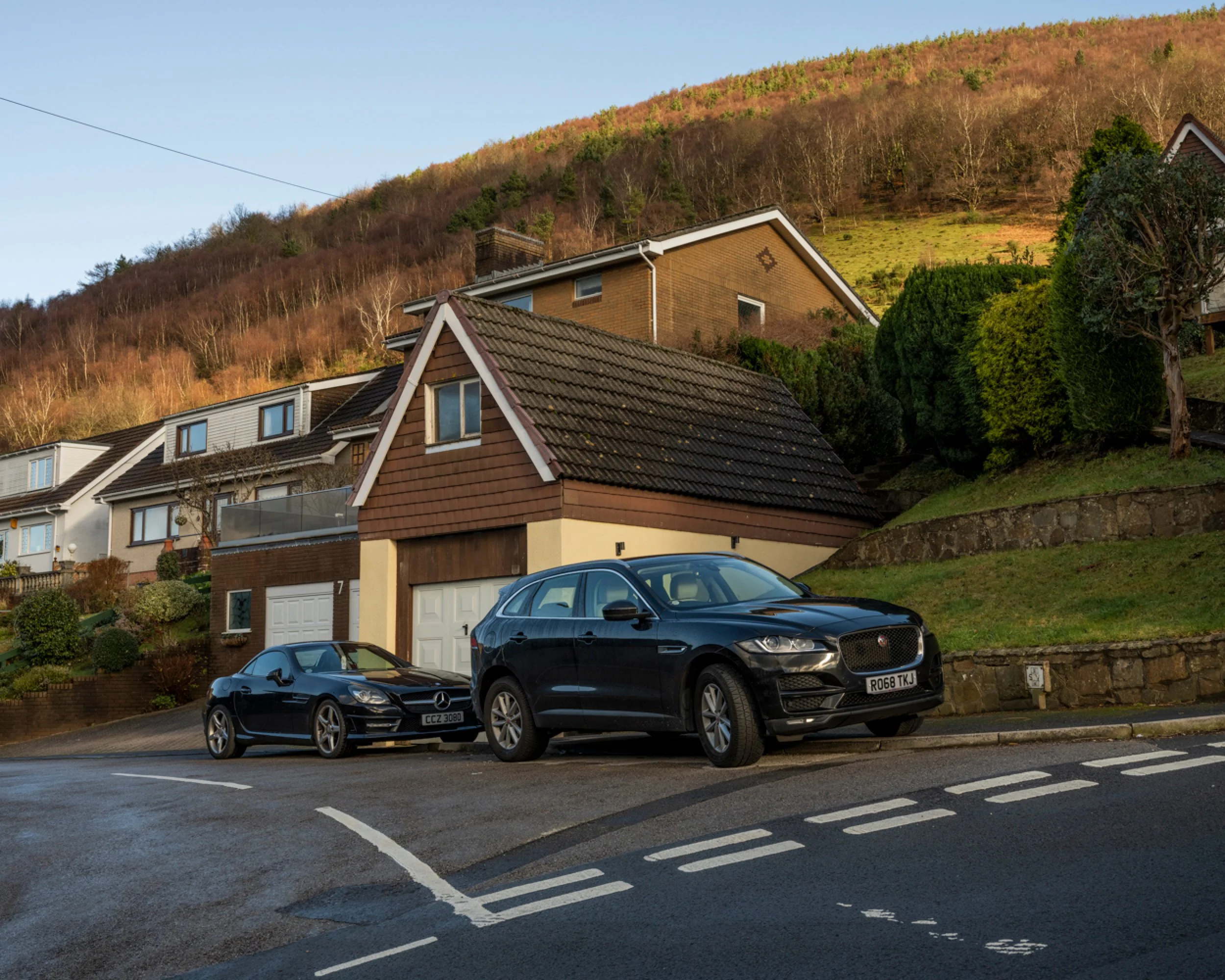 Two black cars parked on a street in front of houses with a hillside in the background.