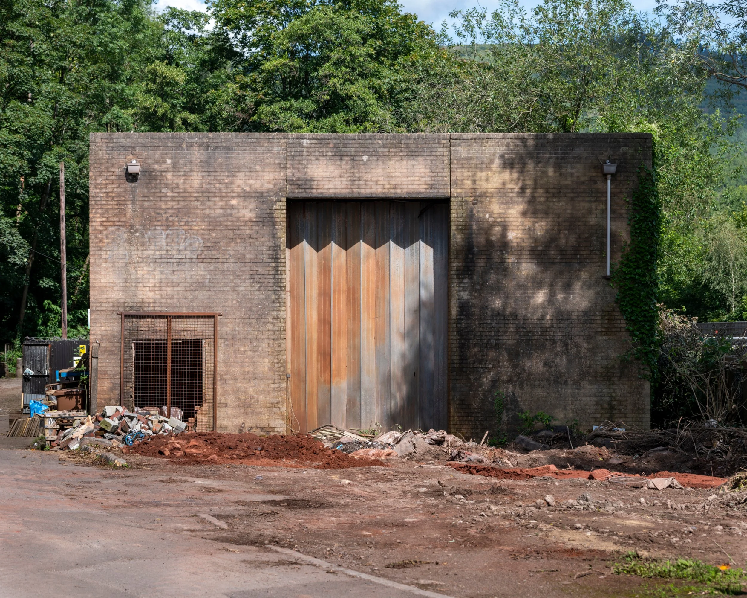 An old brick building with a large wooden gate and a smaller vent door, surrounded by construction debris and greenery.