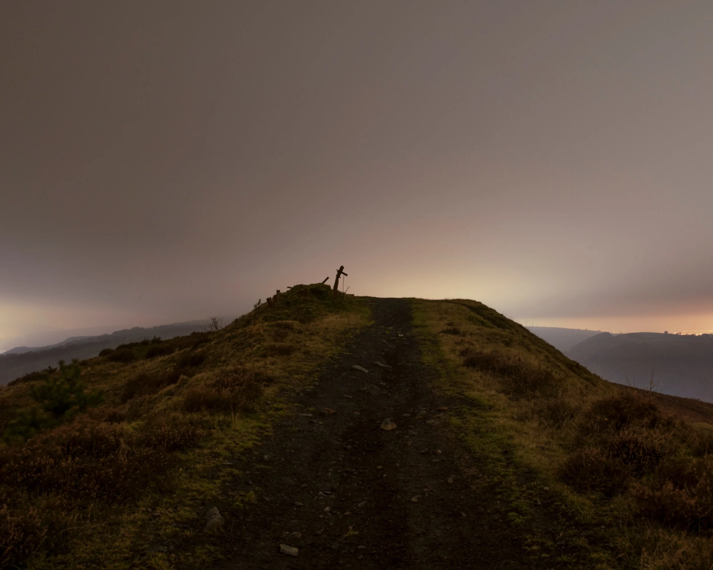 A dirt trail leading up a grassy hill with two wooden crosses at the top, under a cloudy sky at dusk or dawn.