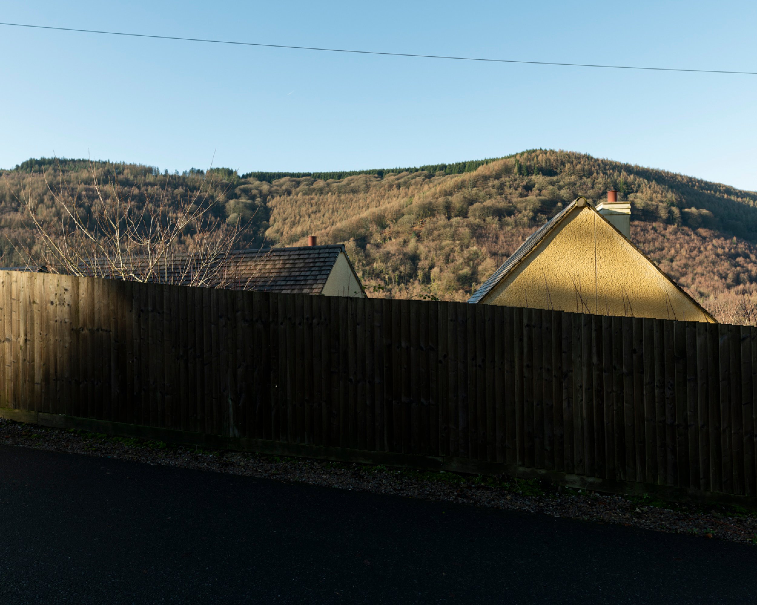 Hilly landscape with trees, two houses with sloped roofs, and a dark wooden fence in the foreground on a clear day.