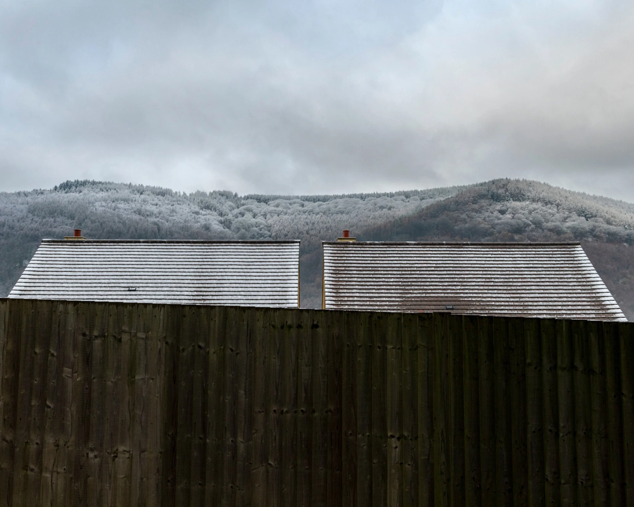 Two houses with snow-covered roofs behind a tall wooden fence, with snow-capped hills and overcast sky in the background.