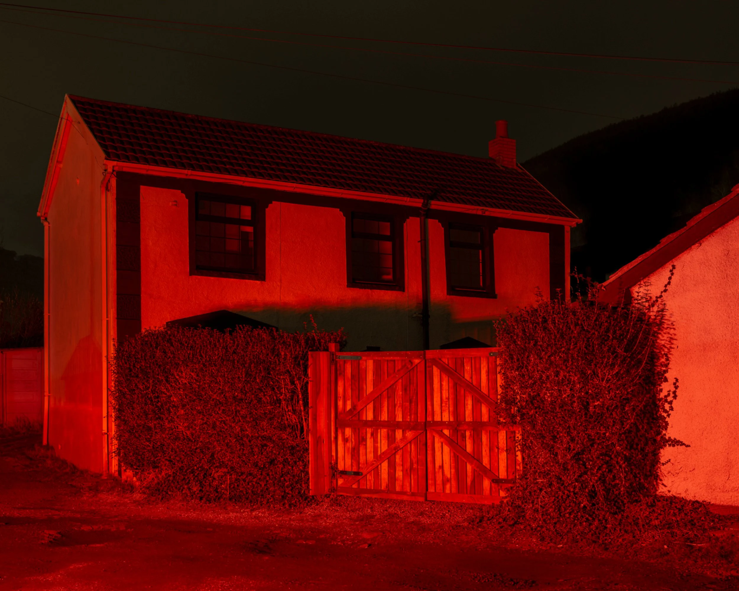 A two-story house illuminated by red light at night with a wooden gate and bushes in front.