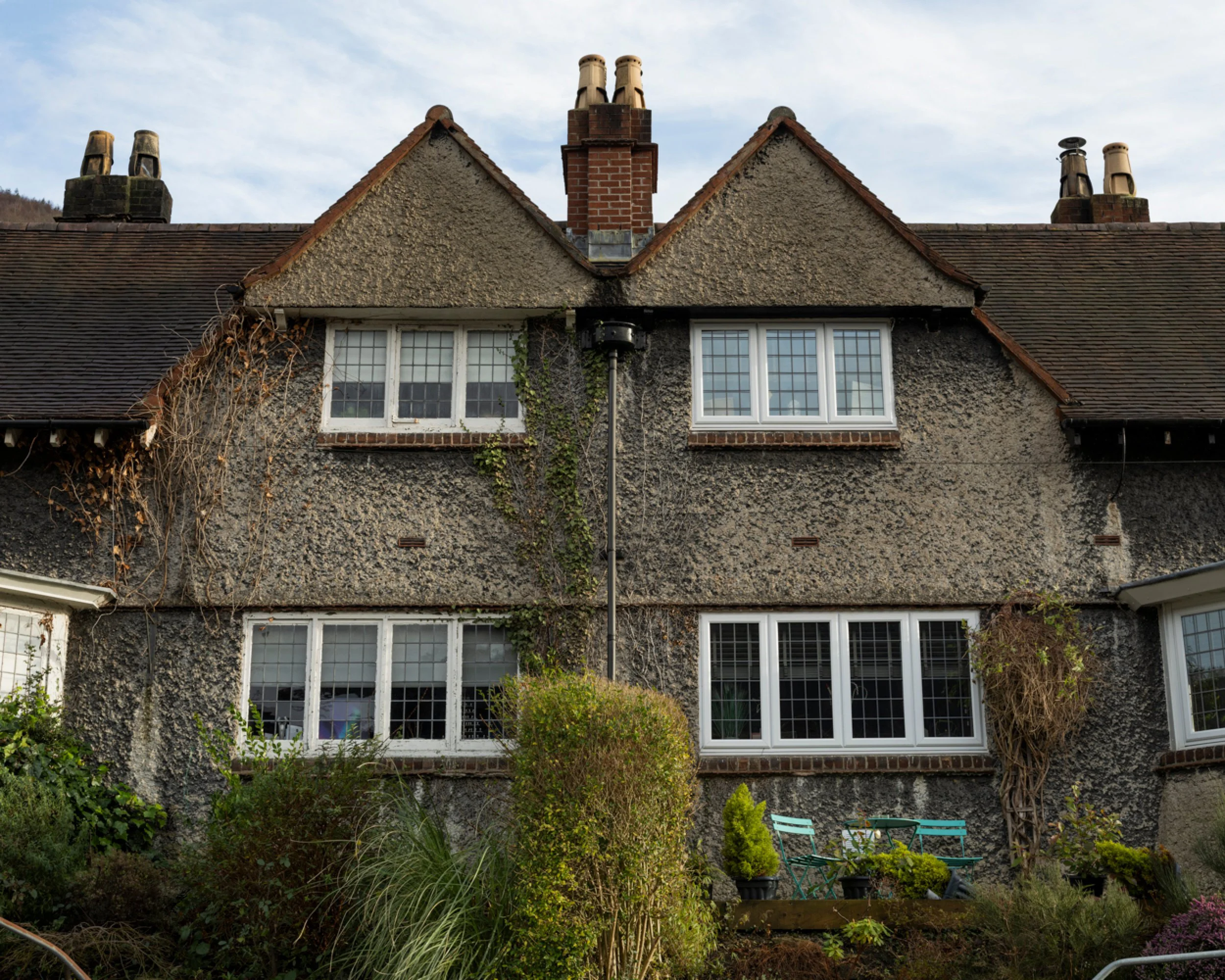 The back of a two-story house with a textured brick exterior, multiple white-framed windows, and a sloped roof with several chimneys. There is greenery and outdoor furniture in the garden.