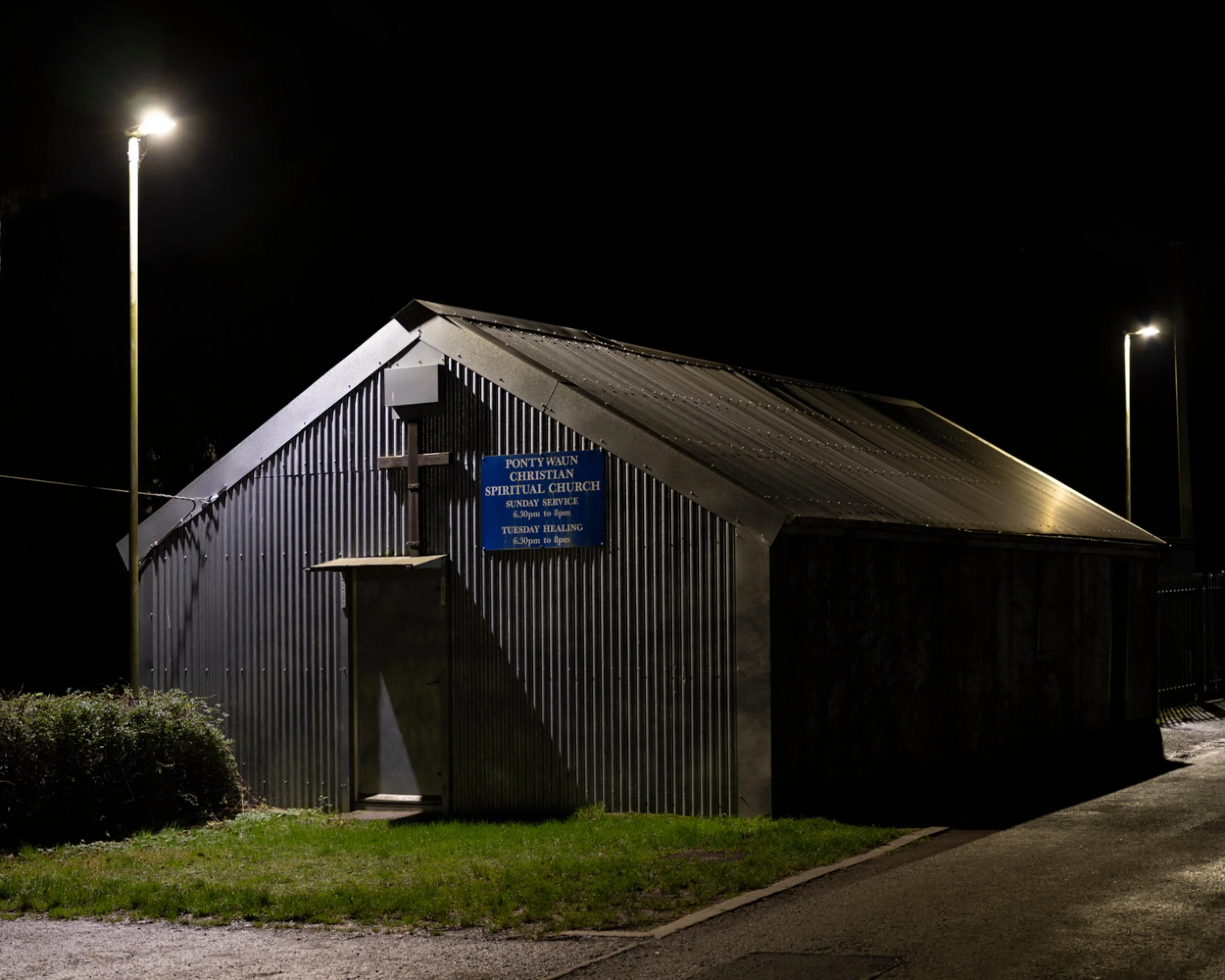 A small church building at night with a sign that reads 'Ponty Waun Christian Spiritual Church'. The church has corrugated metal walls, a cross above the entrance, and is illuminated by streetlights.