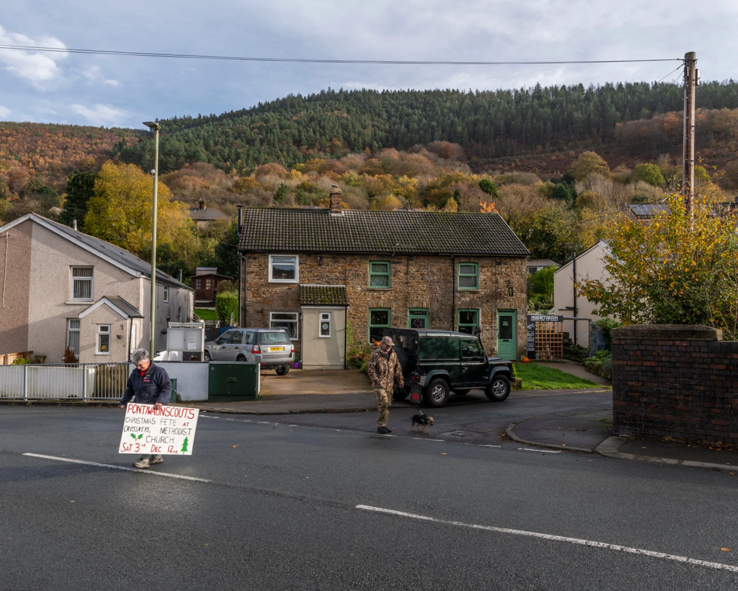 A man holding a sign for scout Christmas fete crossing the street near a black dog with a person in camouflage walking nearby. Behind are houses, parked cars, trees, and a hillside.
