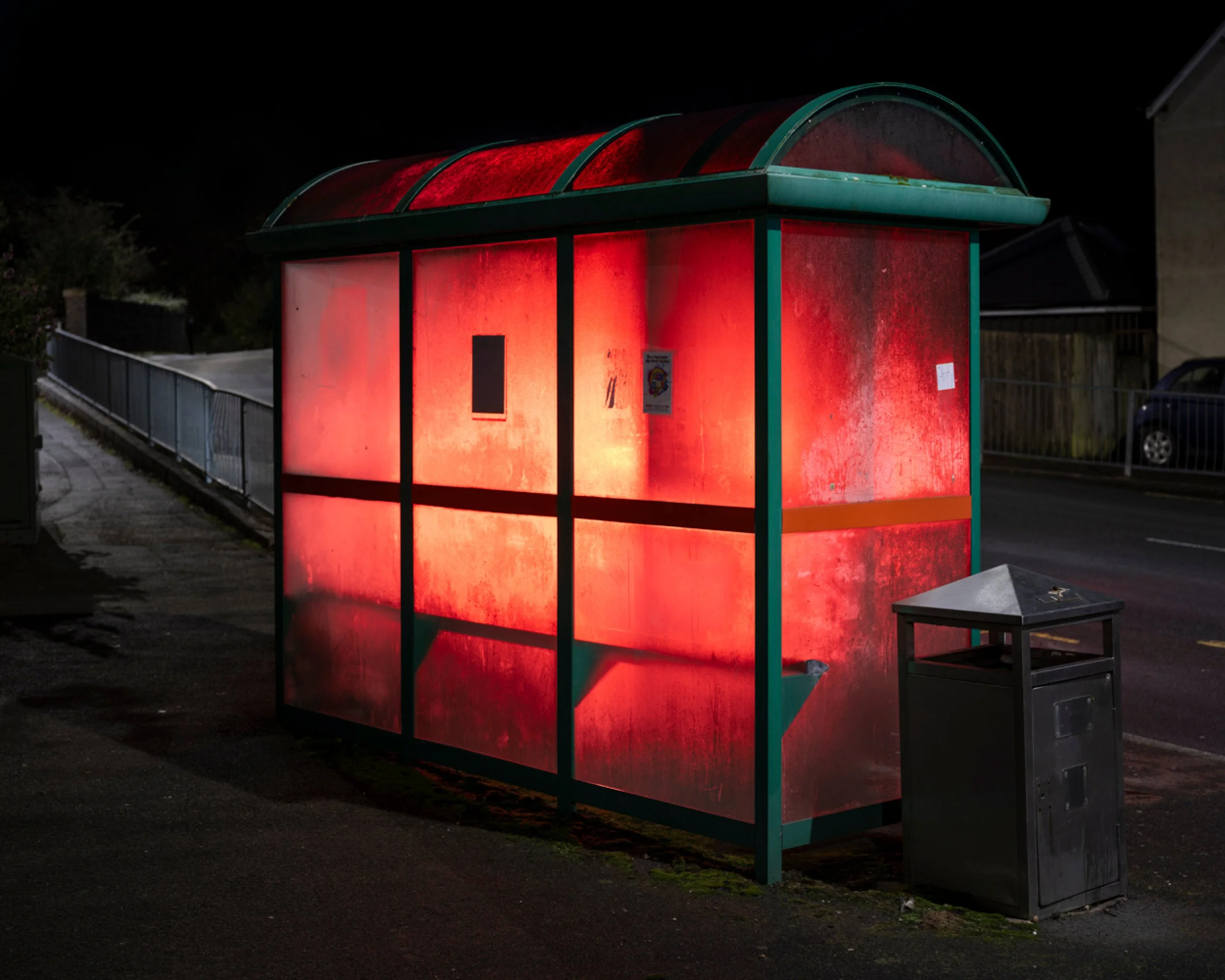 A bus shelter illuminated with red light at night, with a small trash can nearby.