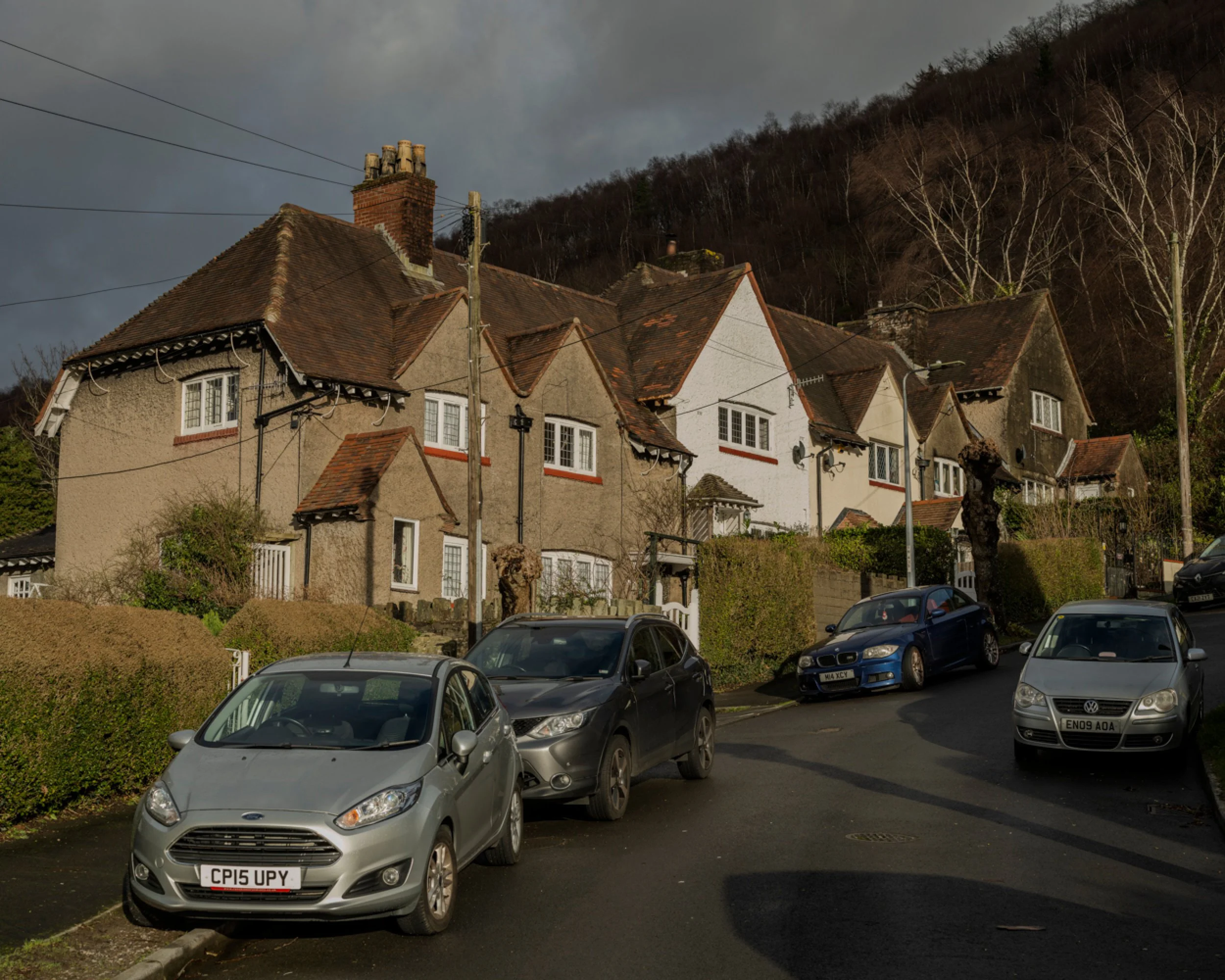 A row of parked cars on a narrow street in front of a hillside neighborhood with old houses that have sloped red-tile roofs and textured stucco exteriors. The sky is cloudy and the hillside is covered with leafless trees.