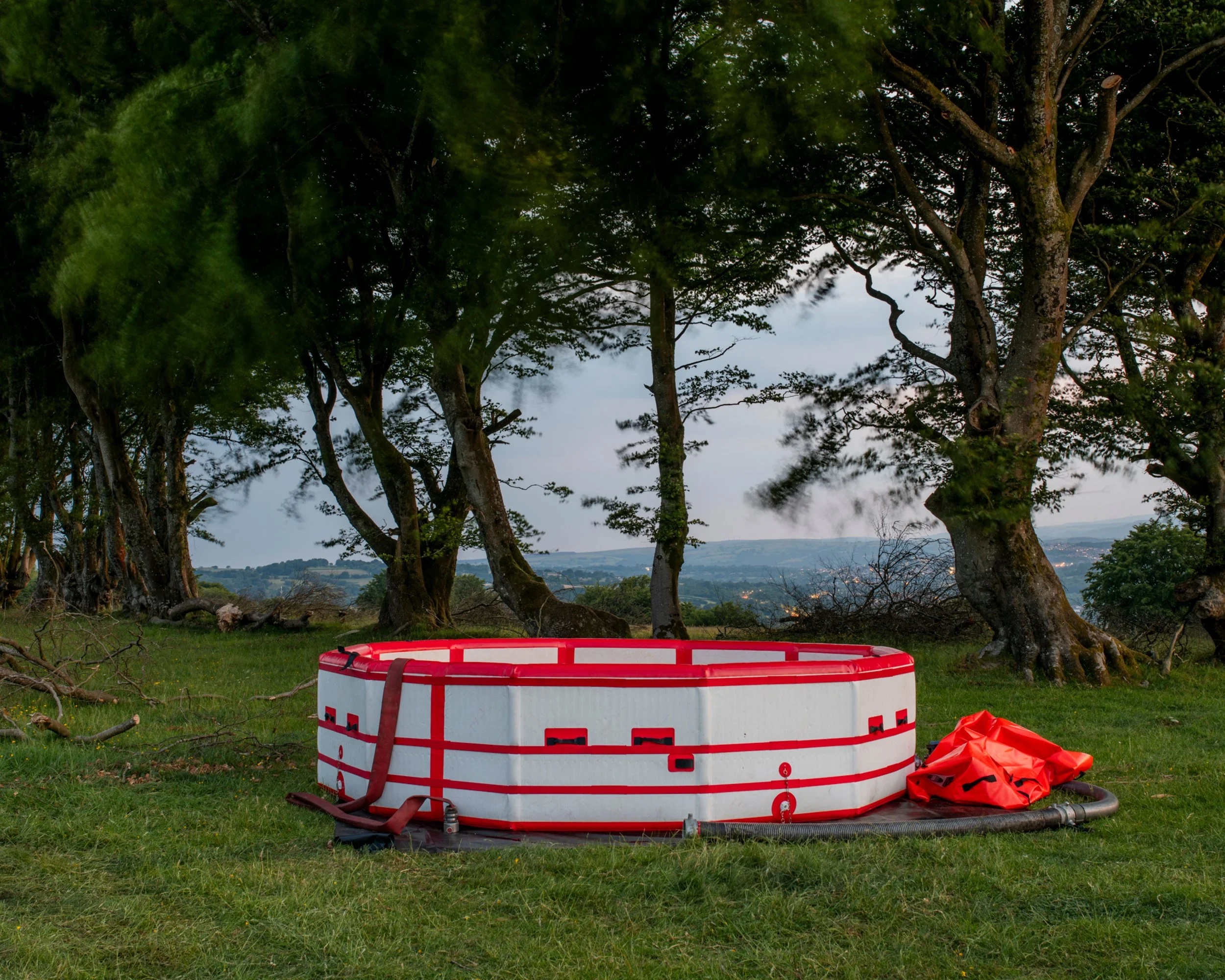 A red and white floating hot tub or fish pond on a grassy area outdoors, with trees in the background and a cloudy sky.