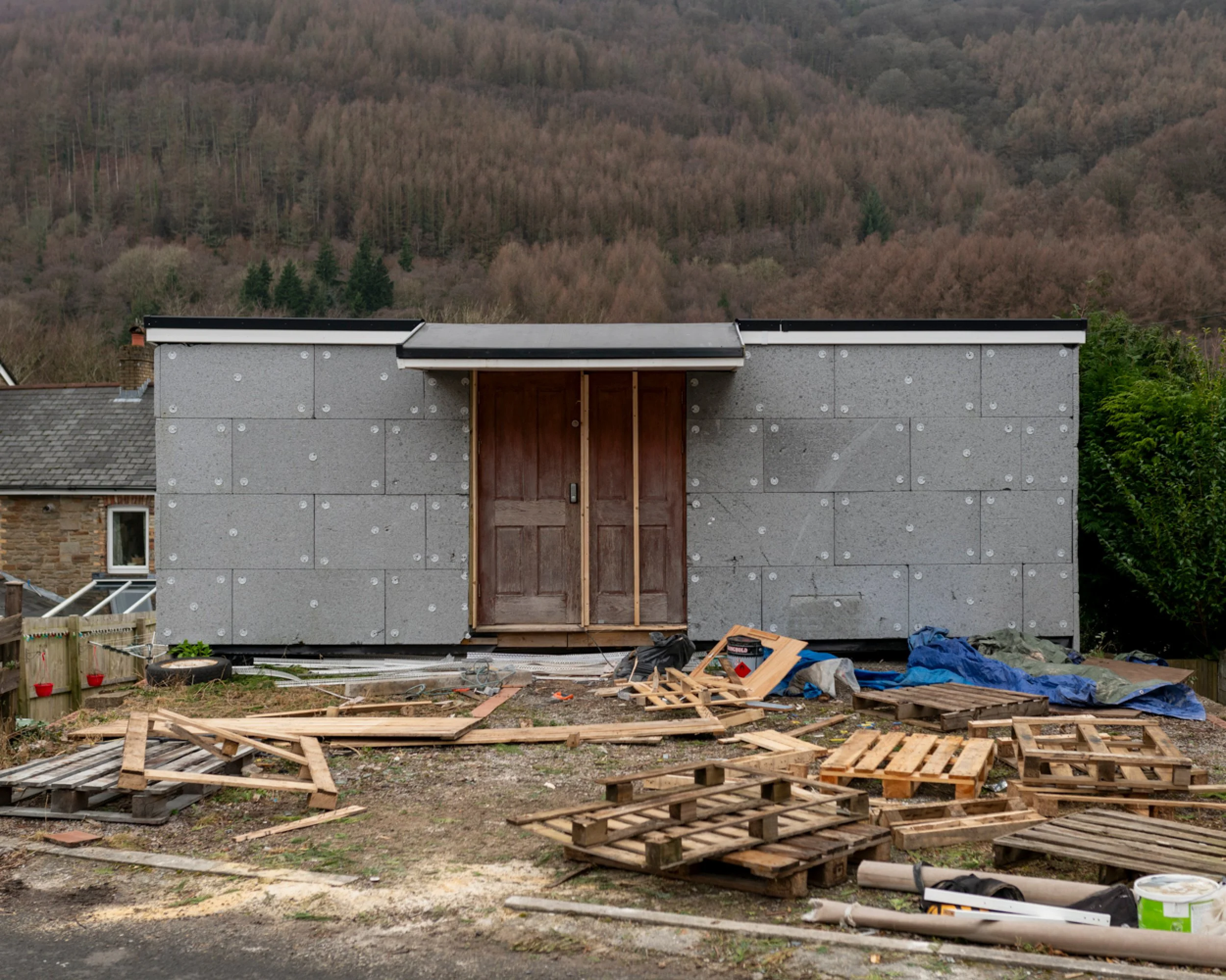 Construction site with a gray building under construction, surrounded by wooden pallets, construction materials, and tools. The building has a wooden door and is set against a backdrop of hills with trees.