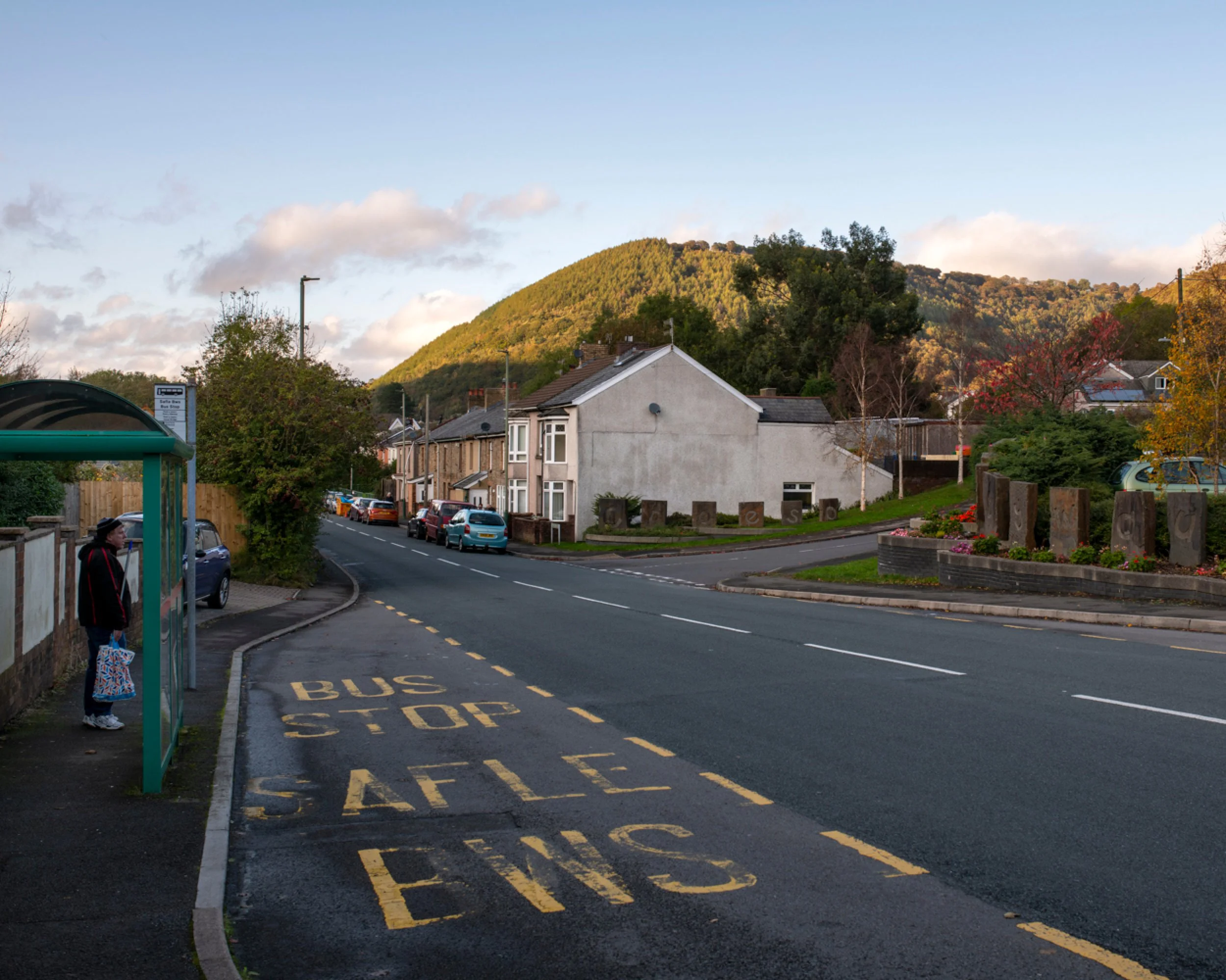 A woman at a bus stop on a residential street, with a painted message in the bus lane that reads 'BUS STOP SAFE' and a background of houses, trees, parked cars, and a hill covered in greenery, during sunset.