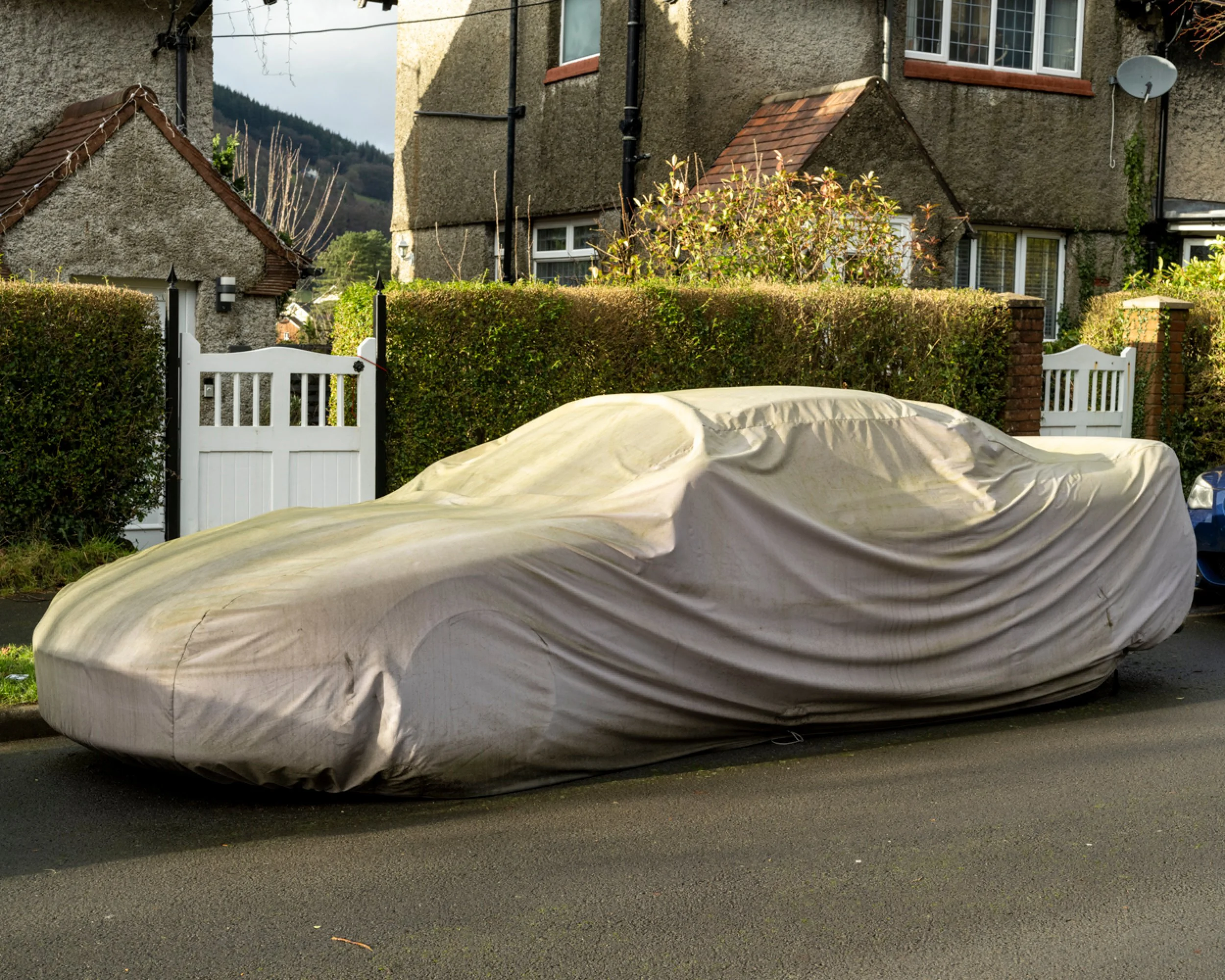 A car covered with a beige protective cover parked on a residential street, with houses, a hedge, and a wooden gate in the background.