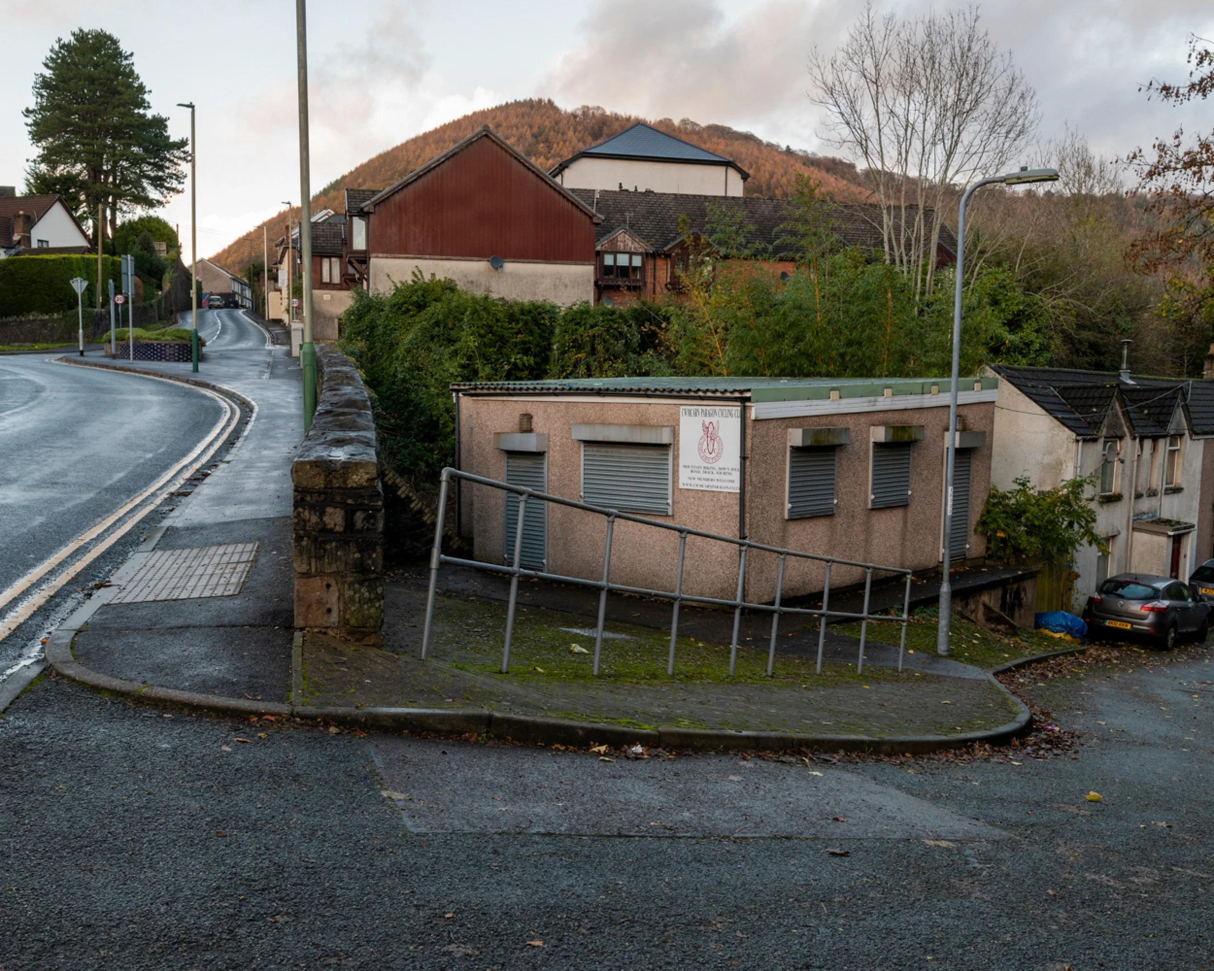 Street view showing a small building with shutters, a railing ramp, parked cars, residential houses, trees, streetlights, and a wooded hill in the background.