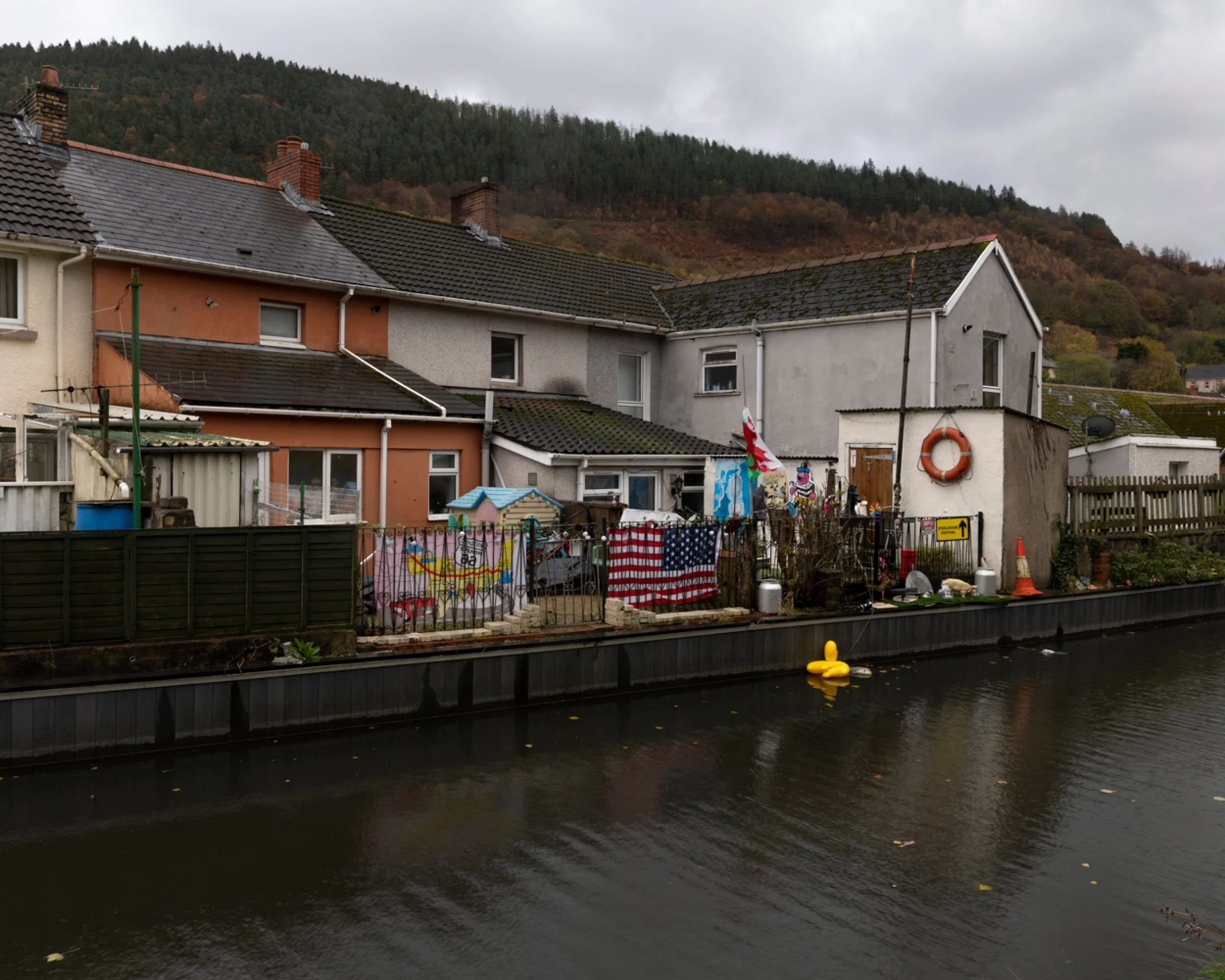 Row of houses along a canal with a grassy hillside and trees in the background. Clothes and flags hang on a fence, including an American flag and others, with a yellow rubber duck floating in the water.