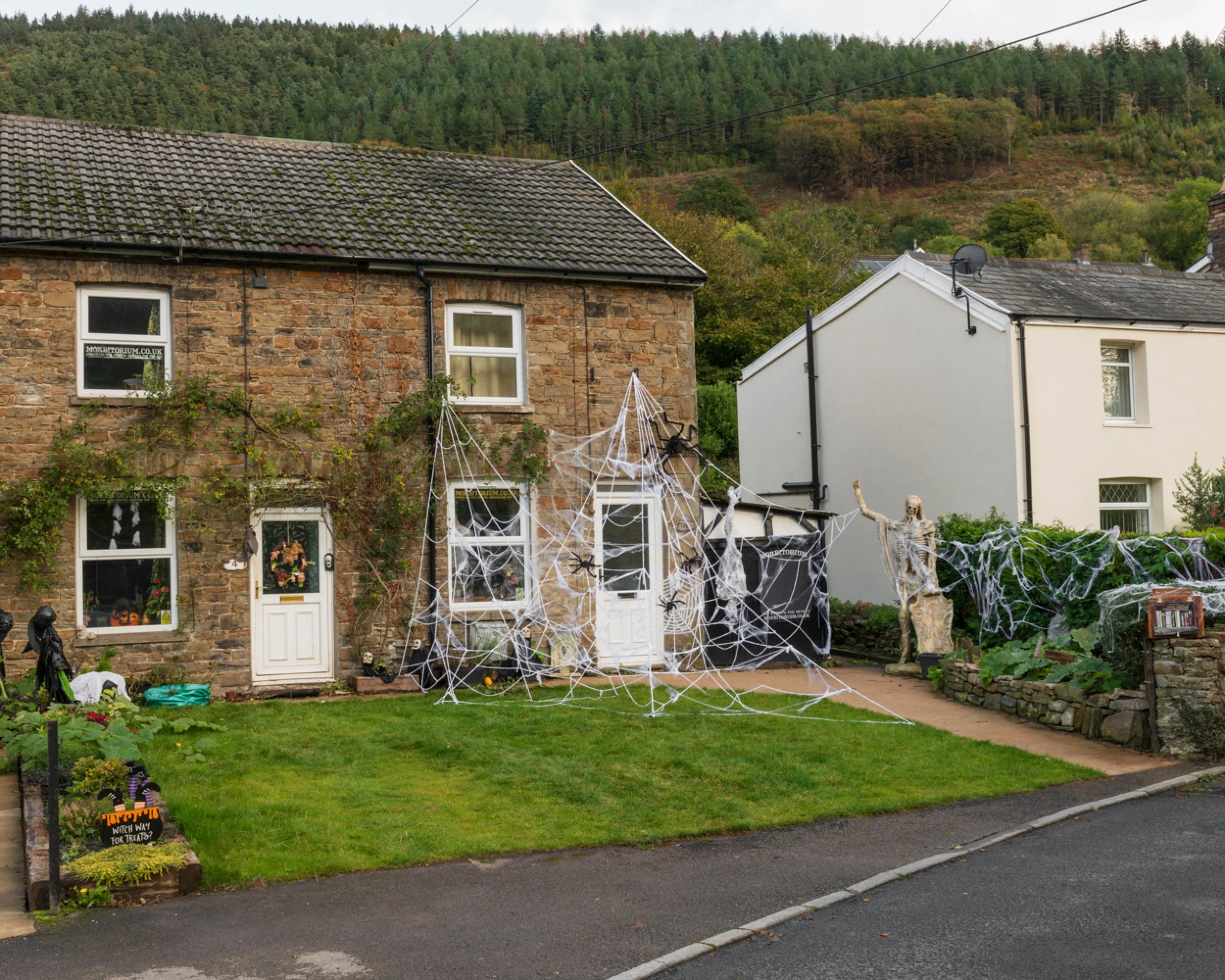 House decorated with Halloween decorations, including fake spider webs, a skeleton figure, and a ghost statue in the yard, suggesting festive Halloween setup.