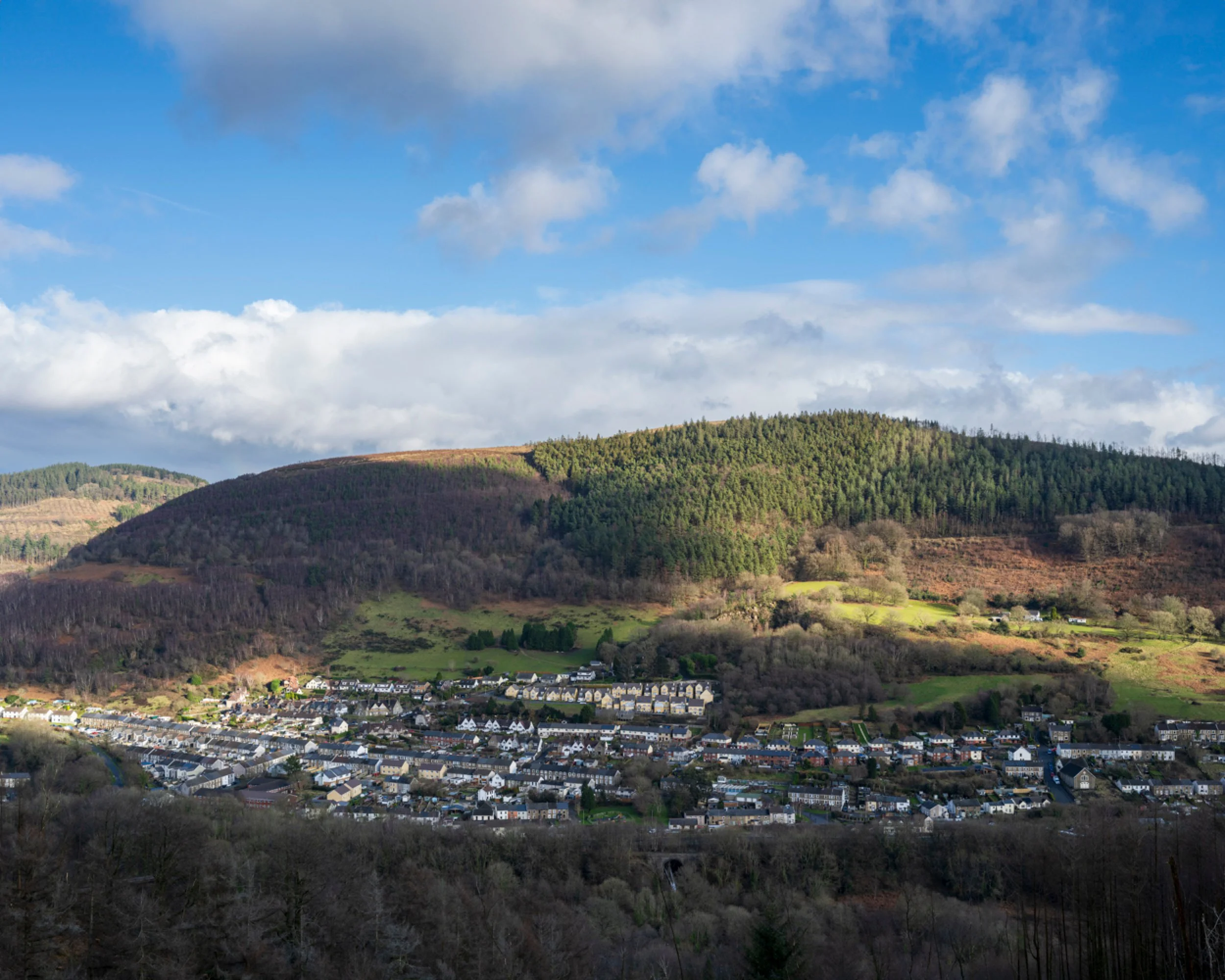 Scenic view of a small town at the base of green hills and mountains under a partly cloudy sky.