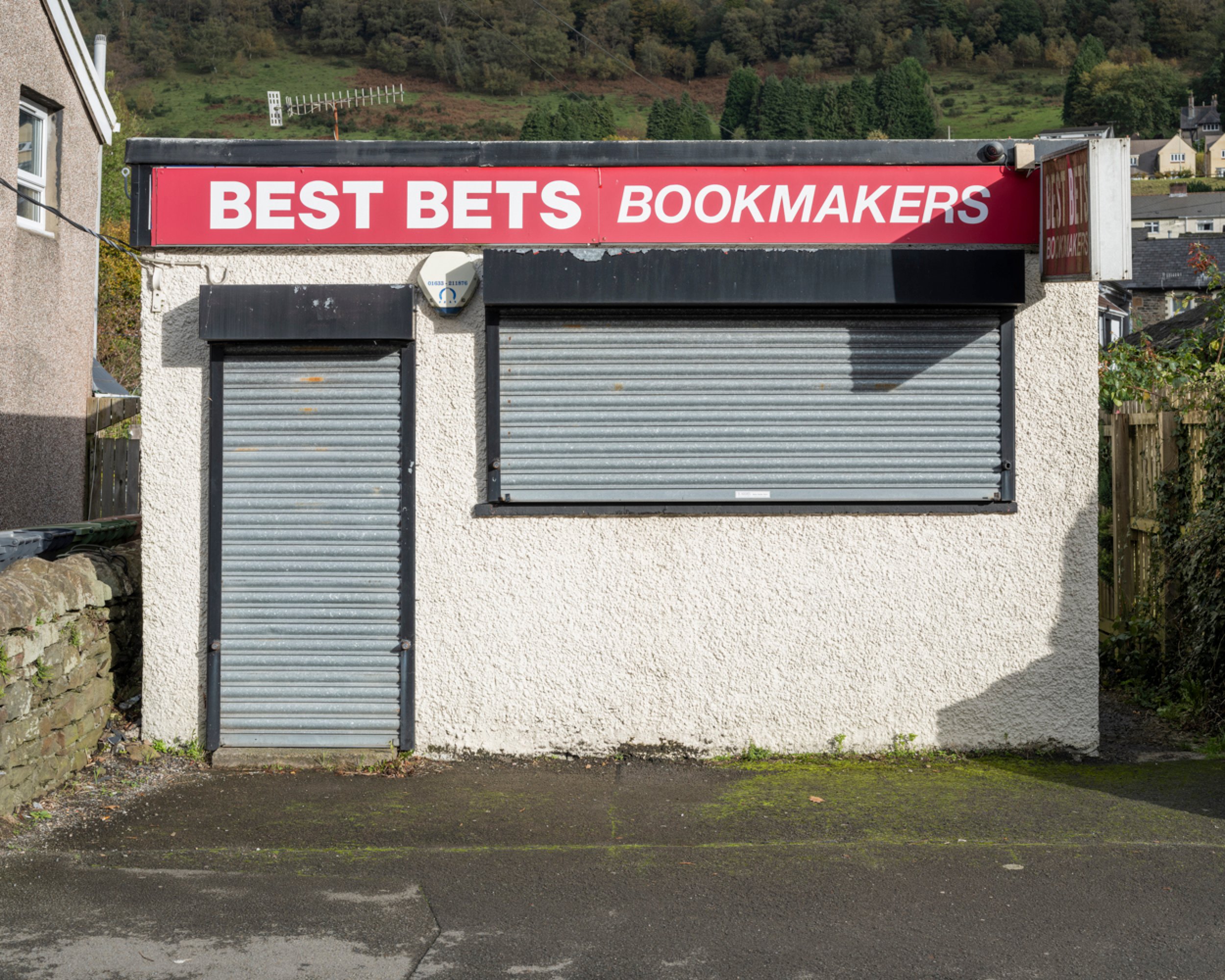 Small building with a red sign reading 'BEST BETS BOOKMAKERS' and closed metal shutters covering windows and door, with a paved sidewalk in the foreground and hillside with trees and houses in the background.