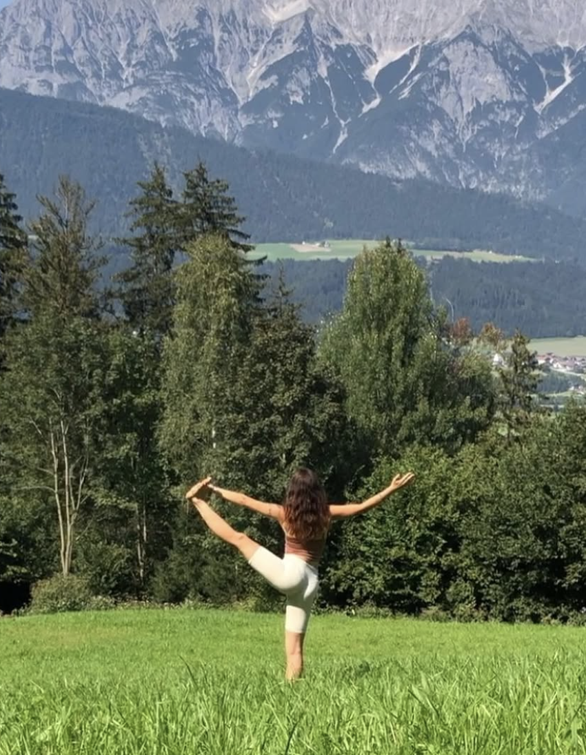 A woman practicing yoga outdoors on grass, balancing on one leg and holding her other leg behind her with one arm, with trees and mountains in the background.