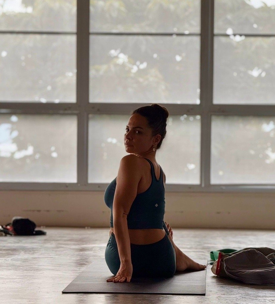 A woman with dark hair in a bun poses in a yoga pose on a mat inside a room with large windows showing a cloudy sky outside.
