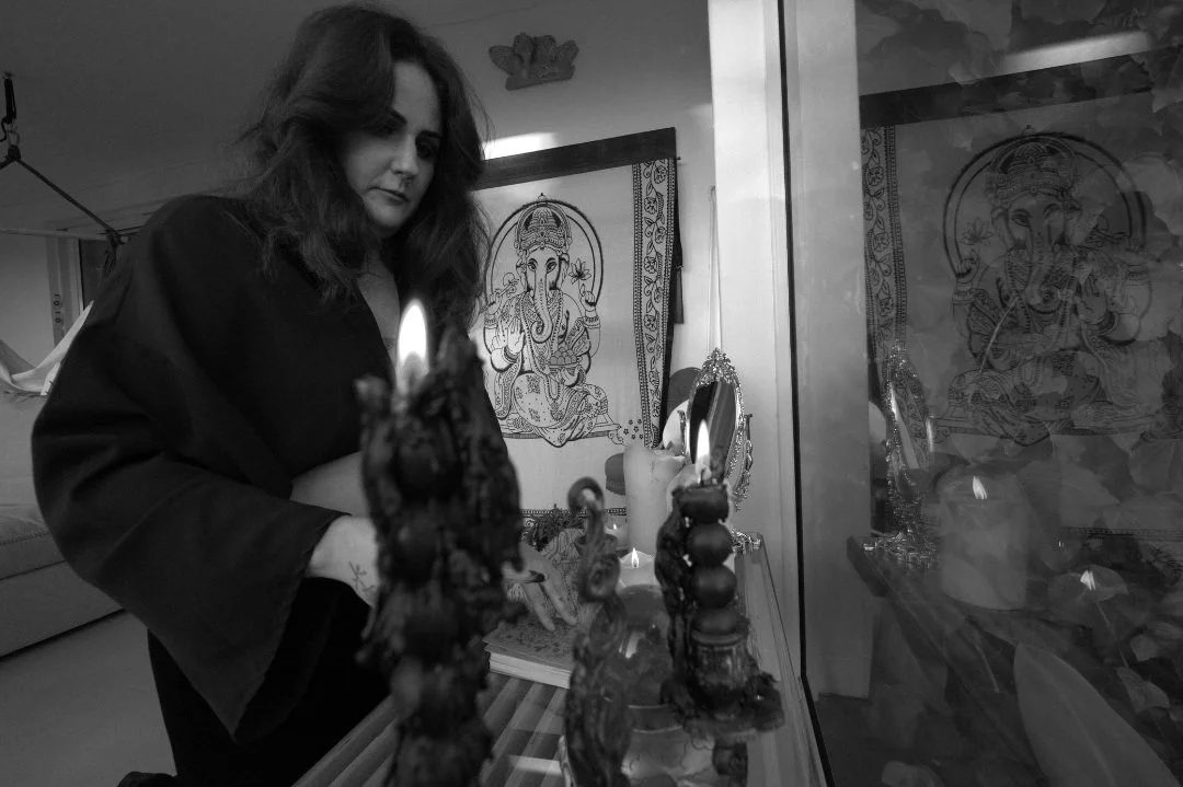 A woman with long, dark hair and a dark top looks at a lit candle in an altar with religious images and decorations, including framed pictures of Ganesha, in a dimly lit room.