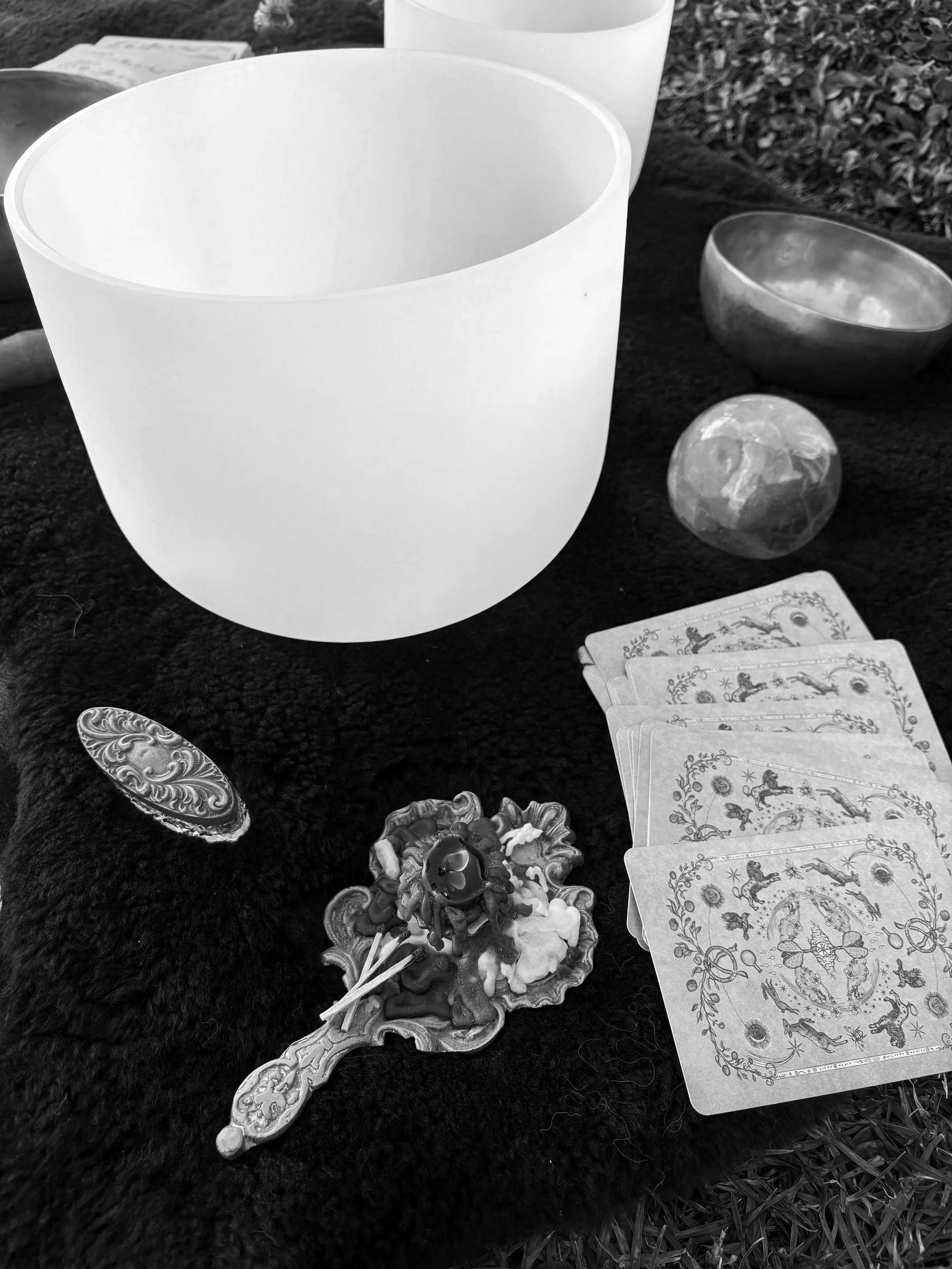 Black and white photo of a crystal singing bowl, a metallic bowl, a small moon-shaped object, a deck of tarot cards, a decorative spoon, and candles placed on a dark soft cloth outdoors.