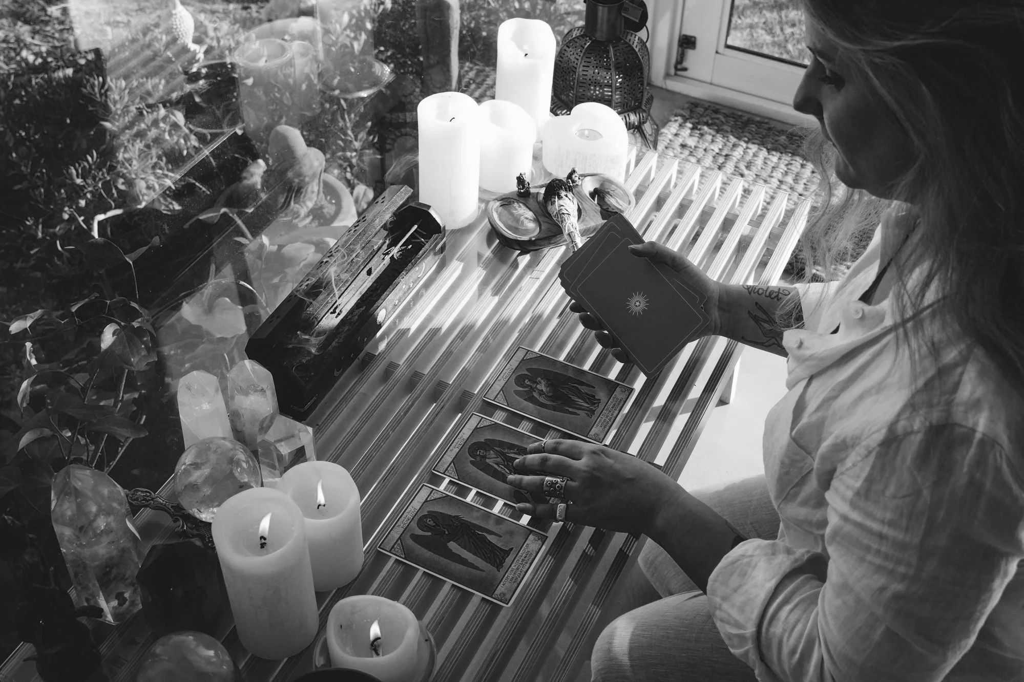 A woman with tattoos and layered jewelry performs a tarot card reading at a table, surrounded by lit candles, crystals, and tarot cards, with a window reflecting trees outside.