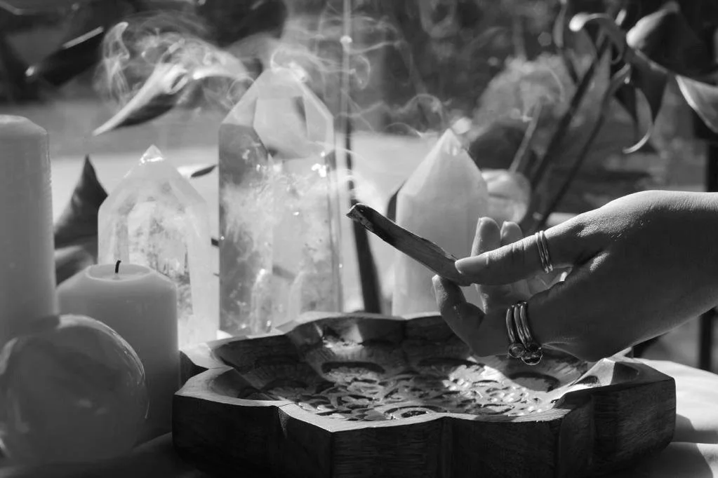 A hand with rings holding a salvia leaf over a wooden bowl, with large crystals, candles, an apple, and other items on a table.