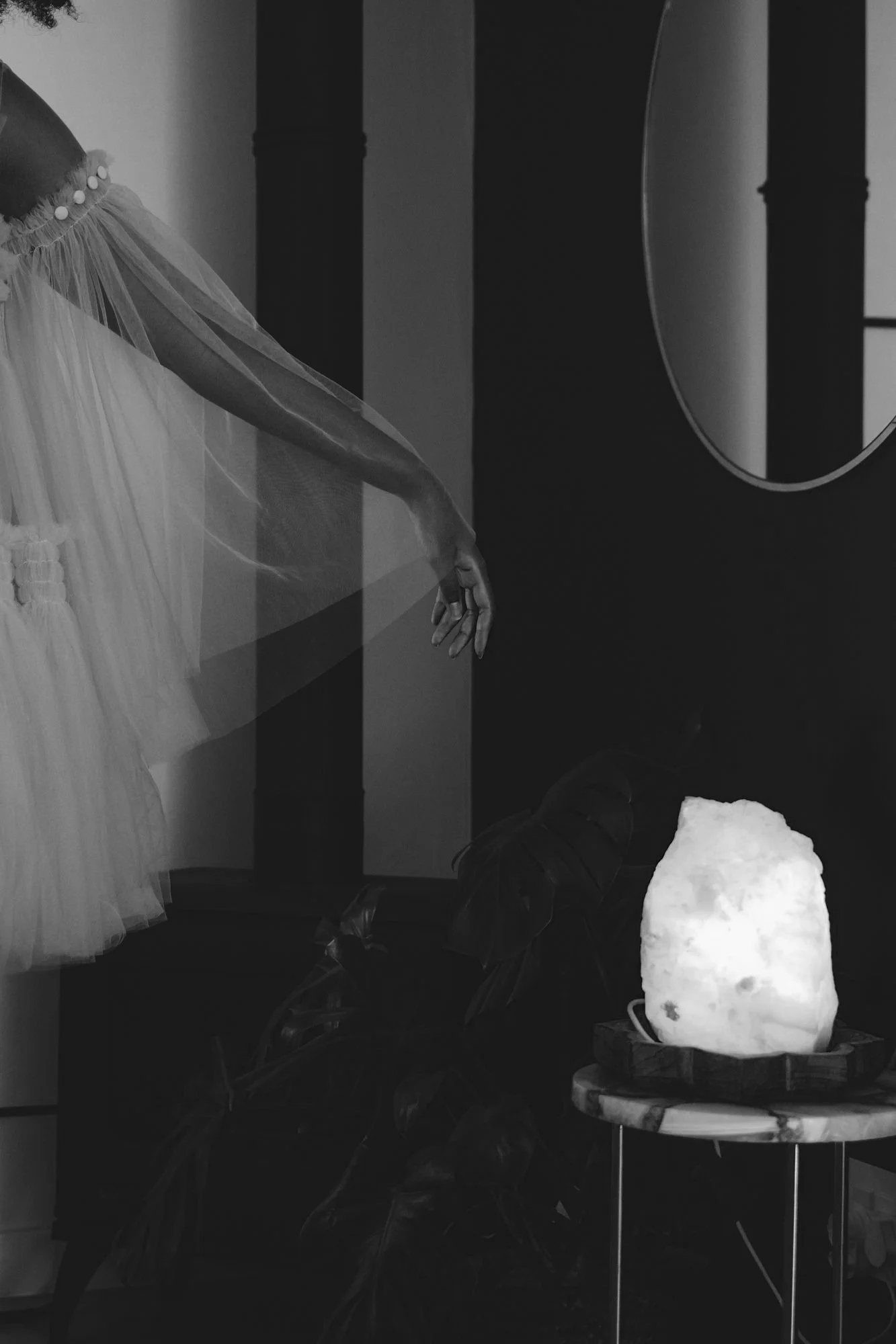 Part of a person wearing a light-colored dress and tulle veil, reaching towards a glowing salt lamp on a small table, with dark interior background and a round mirror on the wall.