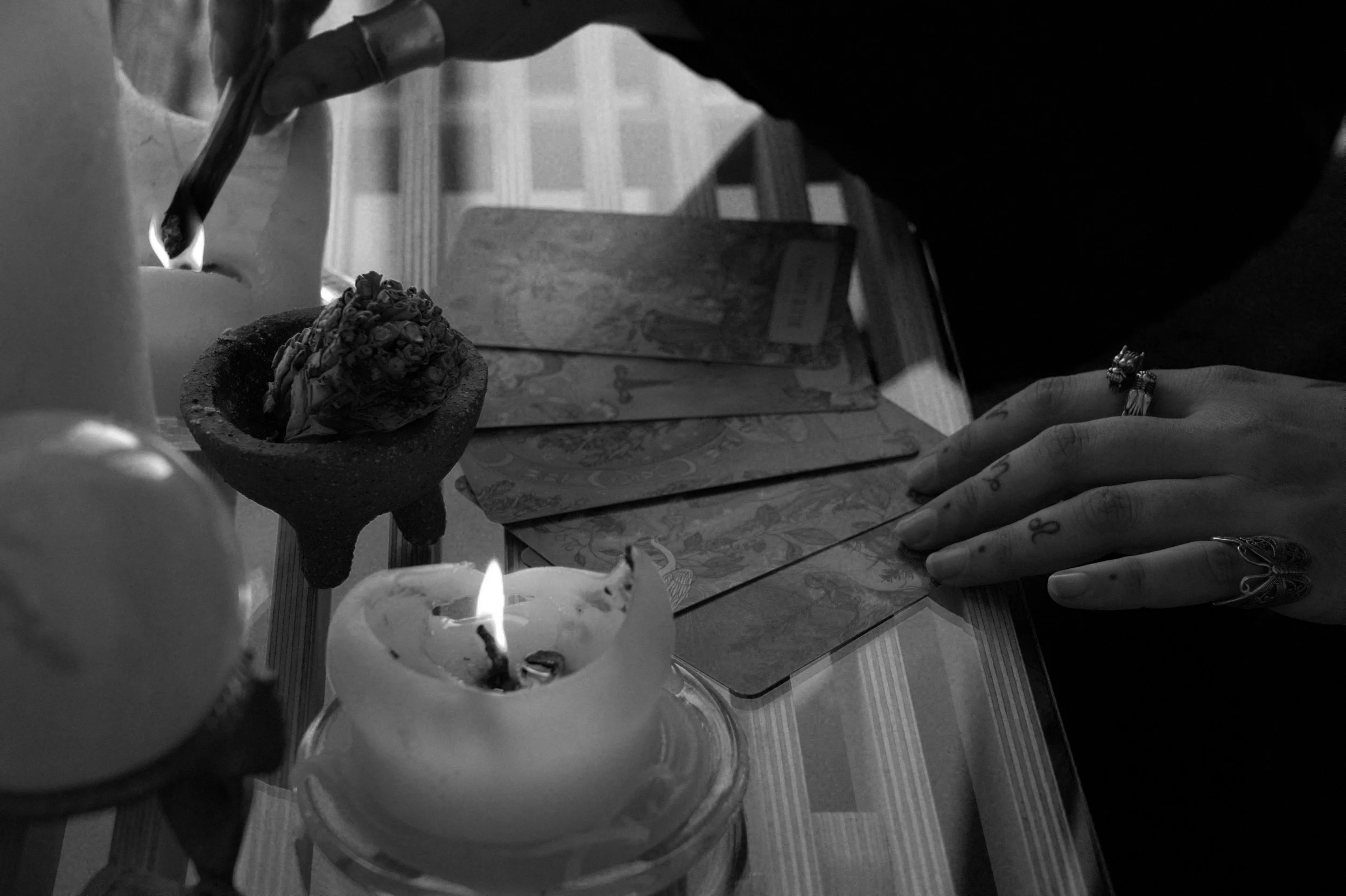 A person is performing a ritual involving burning candles, tarot cards, and an incense cone on a wooden table, with their hands reaching towards the items.