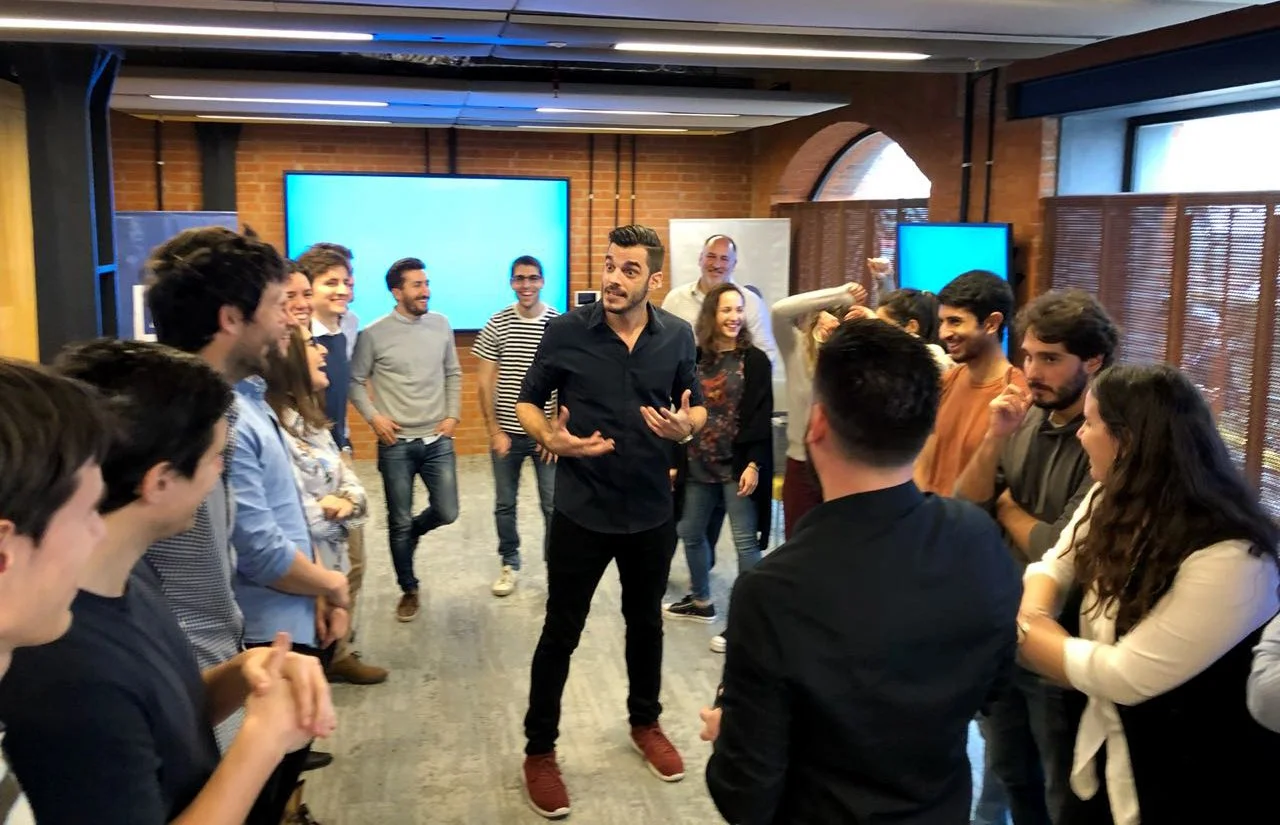 Grupo de personas en una sala de reuniones, algunas ríen y otras escuchan a un hombre que parece estar dando una charla o explicación.