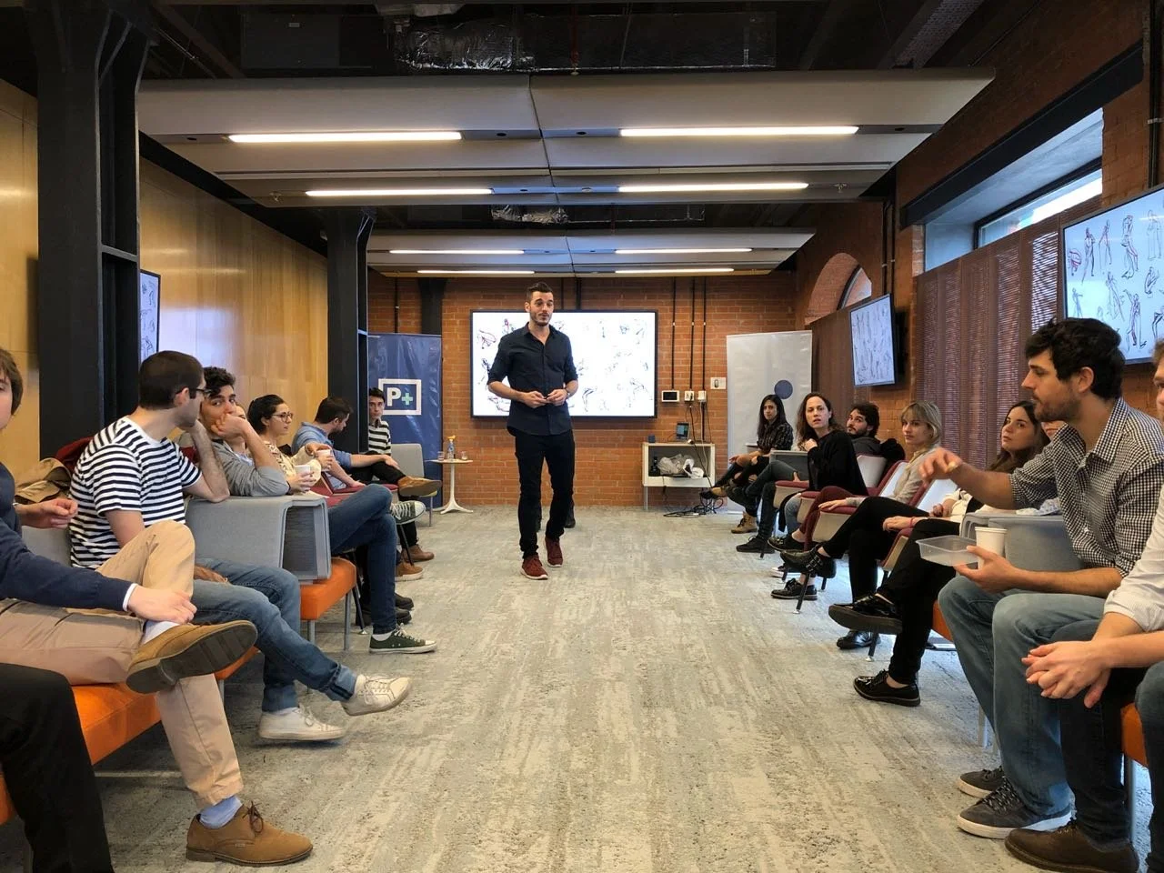 Hombre de pie hablando en una sala de reuniones con personas sentadas a ambos lados de la sala, varias pantallas en las paredes mostrando ilustraciones.