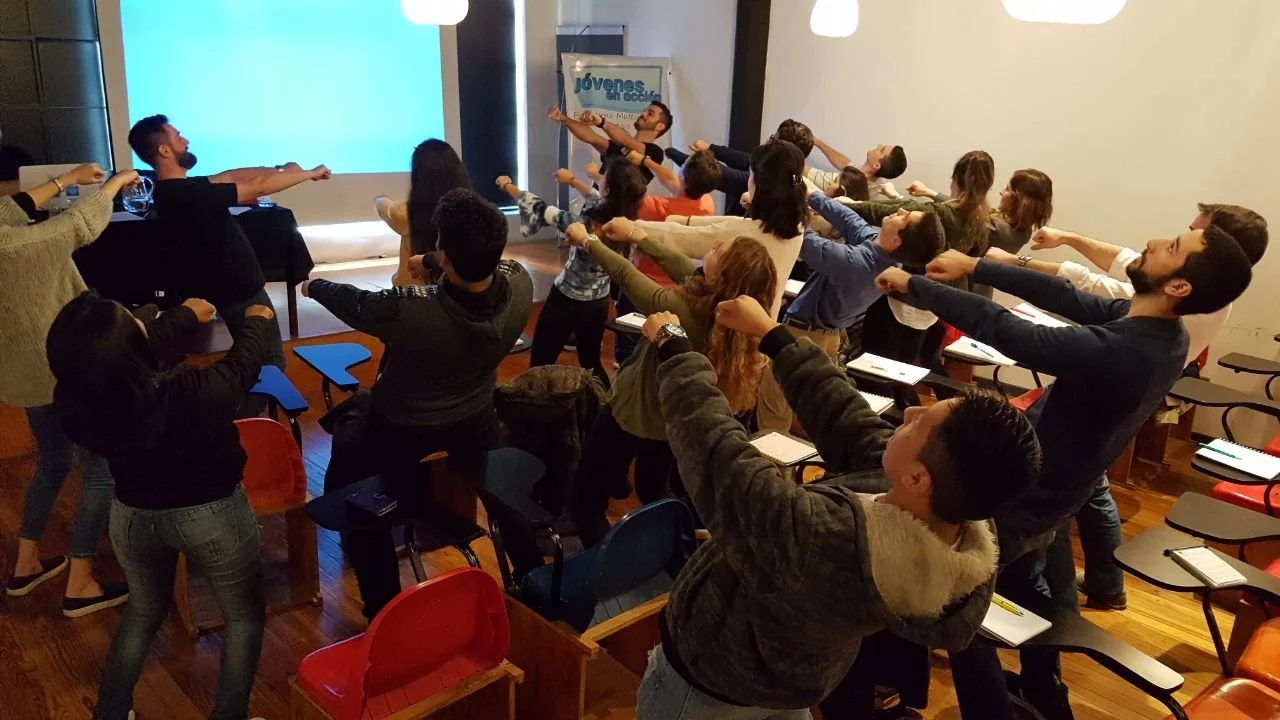 Grupo de personas en una sala participando en una actividad de estiramiento o ejercicio, todos levantando un brazo en línea recta hacia adelante.
