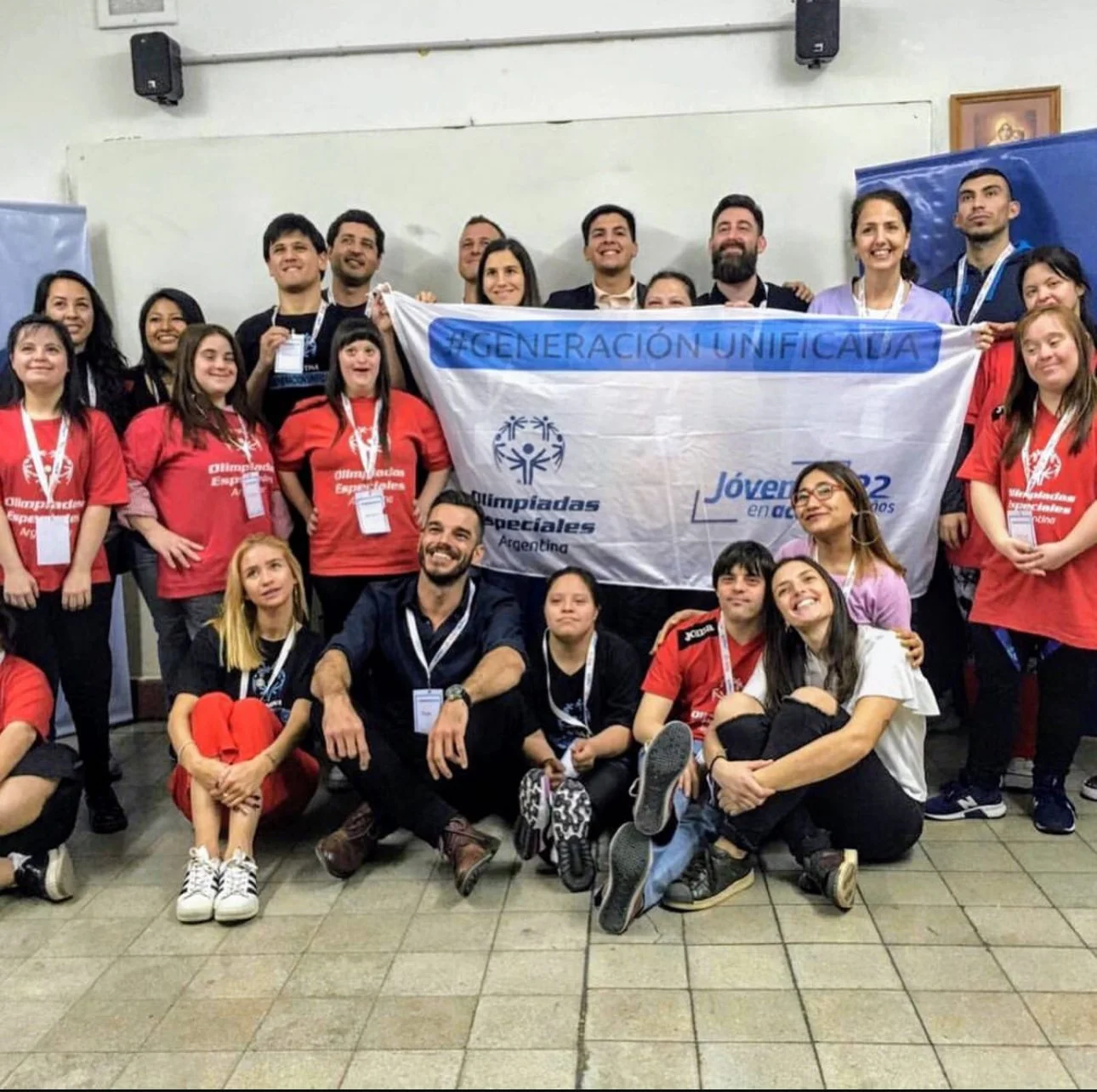 Grupo de personas posando con una bandera que dice #GENERACIÓN UNIFICADA, en un evento de Olimpiadas Especiales en Argentina, algunos llevan camisetas rojas y otros con gafas y medallas