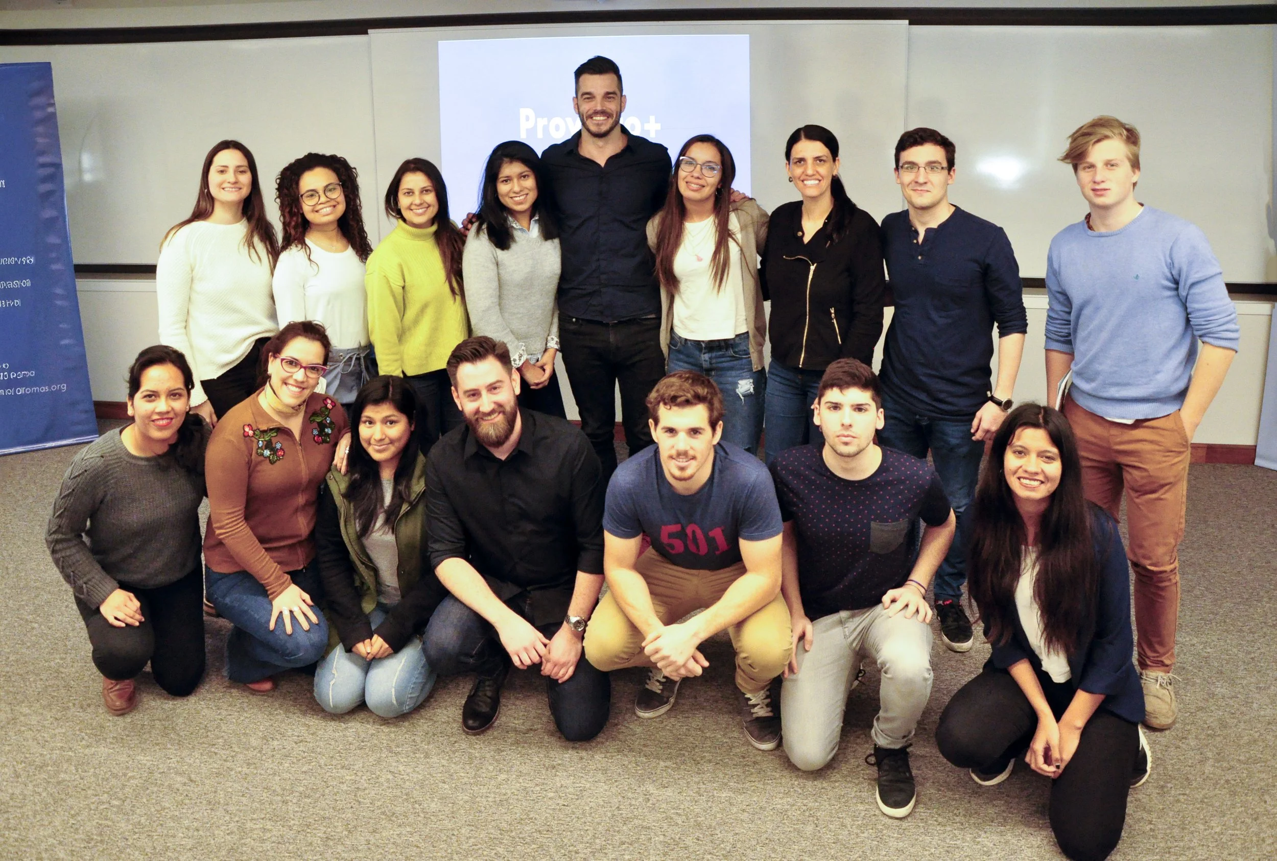 Grupo de personas sonriendo y posando en un salón de clases, con una pantalla en el fondo que dice 'Prov...+'.
