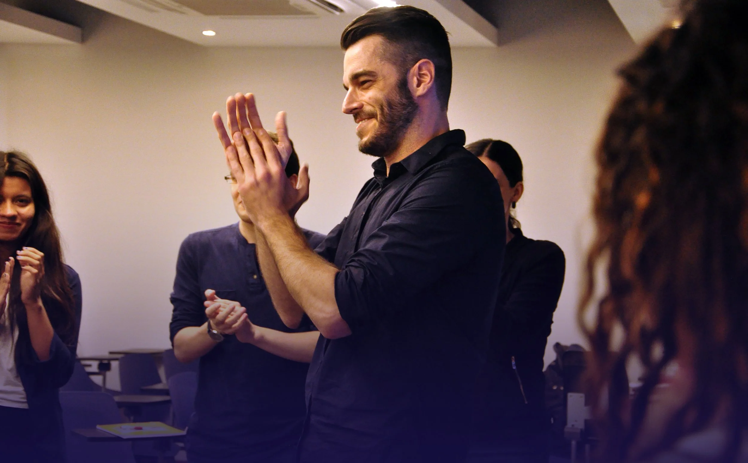 Un hombre sonriendo, sosteniendo sus manos juntas en un gesto de agradecimiento o felicidad, rodeado de varias personas en un ambiente interior, posiblemente en una reunión o celebración.