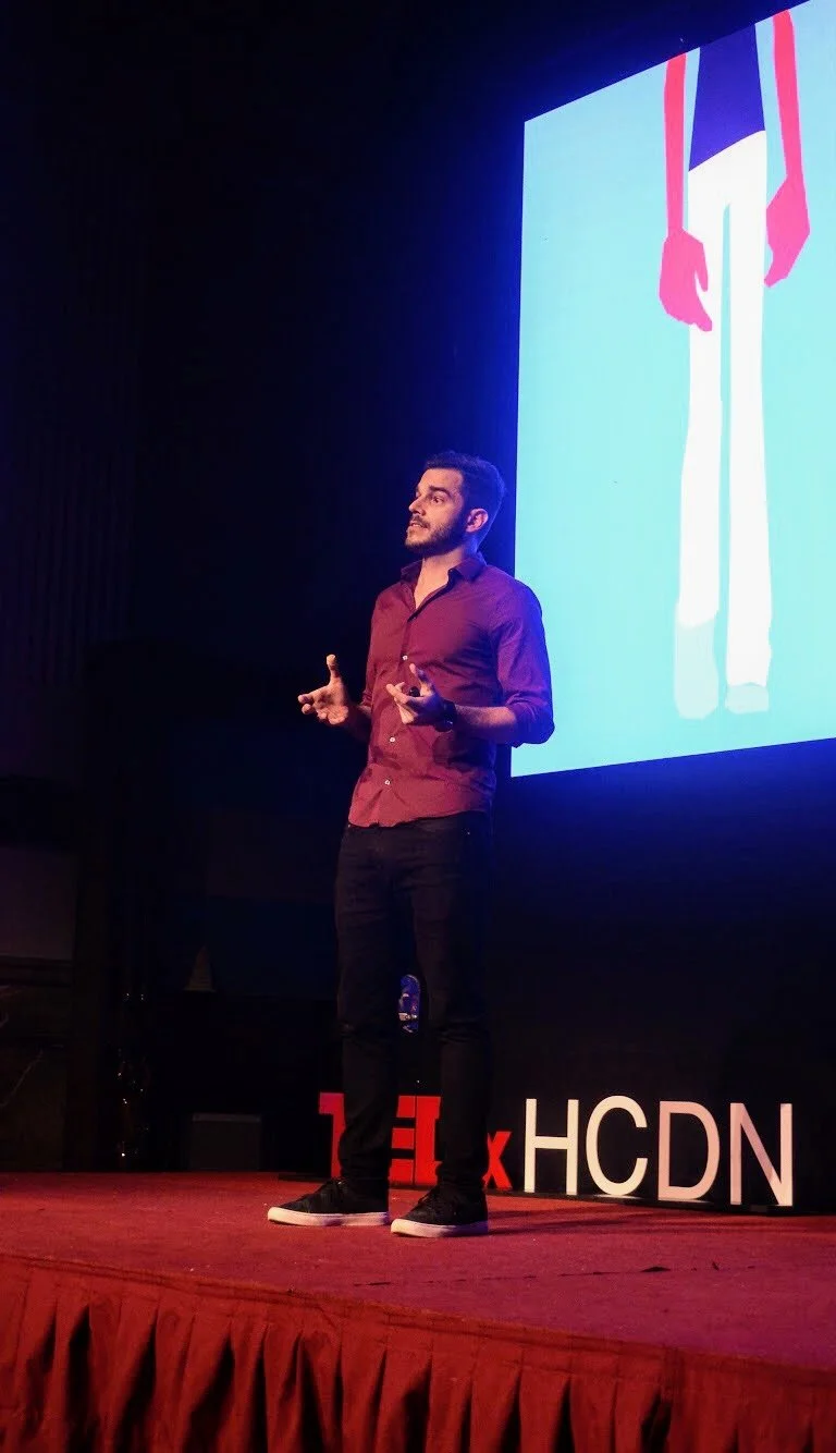 Hombre dando una charla en un escenario con una pantalla grande detrás y letras de TEDx en el suelo.