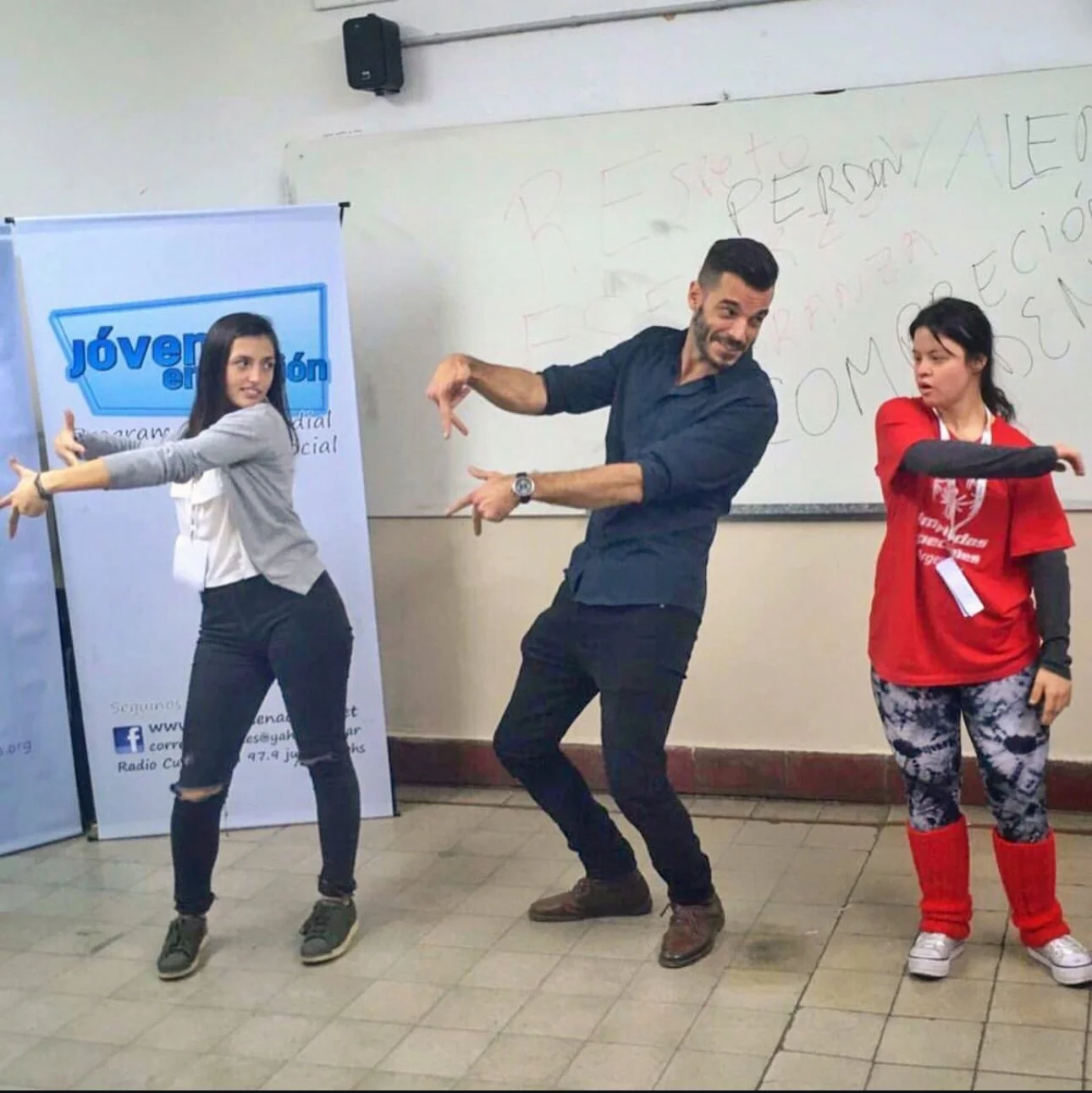 Tres personas, un hombre y dos mujeres, posando como si bailaran en una sala de clases con una pizarra y un cartel.