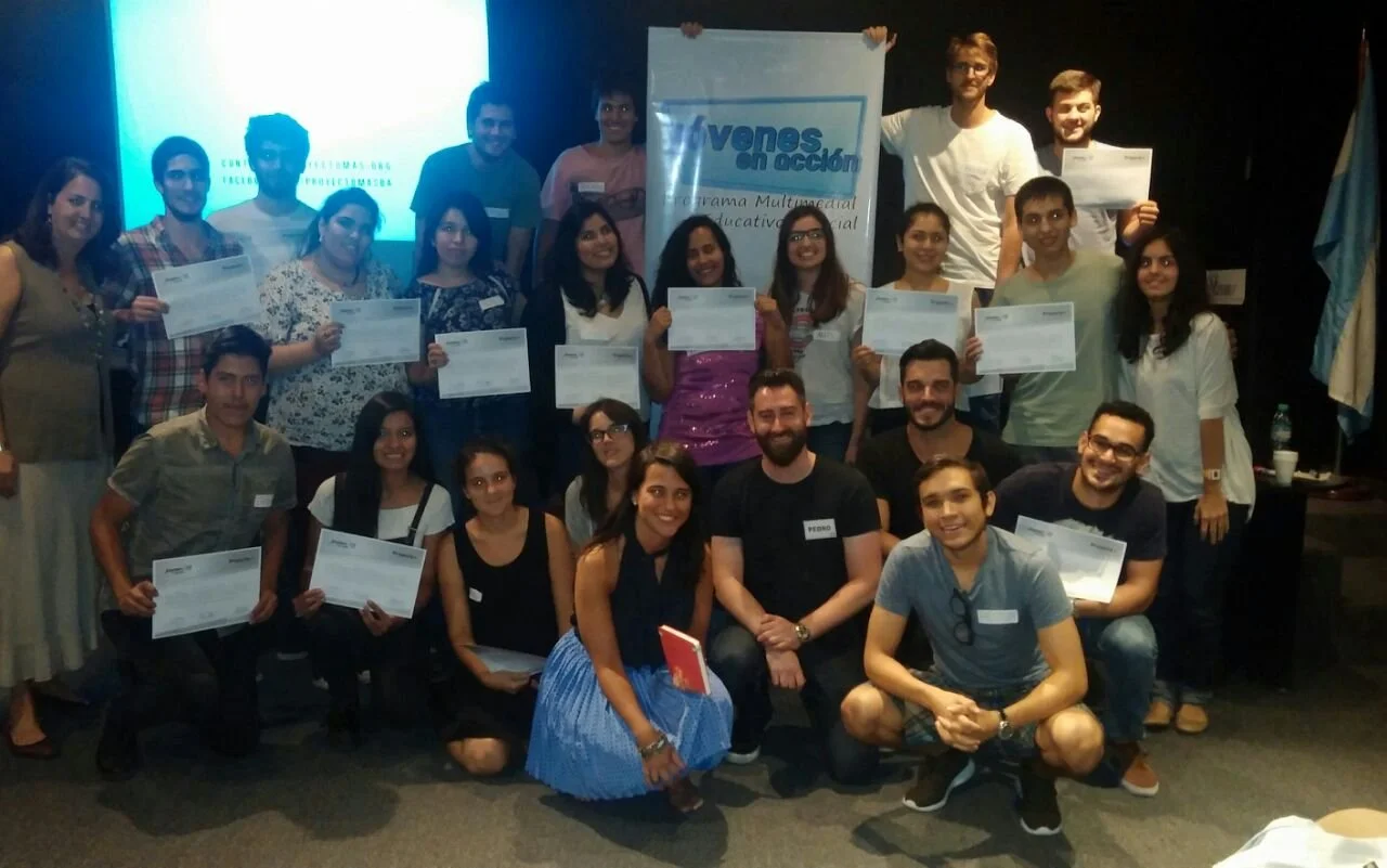 Grupo de personas en una ceremonia de reconocimiento, algunas sosteniendo diplomas, con una bandera y un cartel que dice 'Jóvenes en acción'.