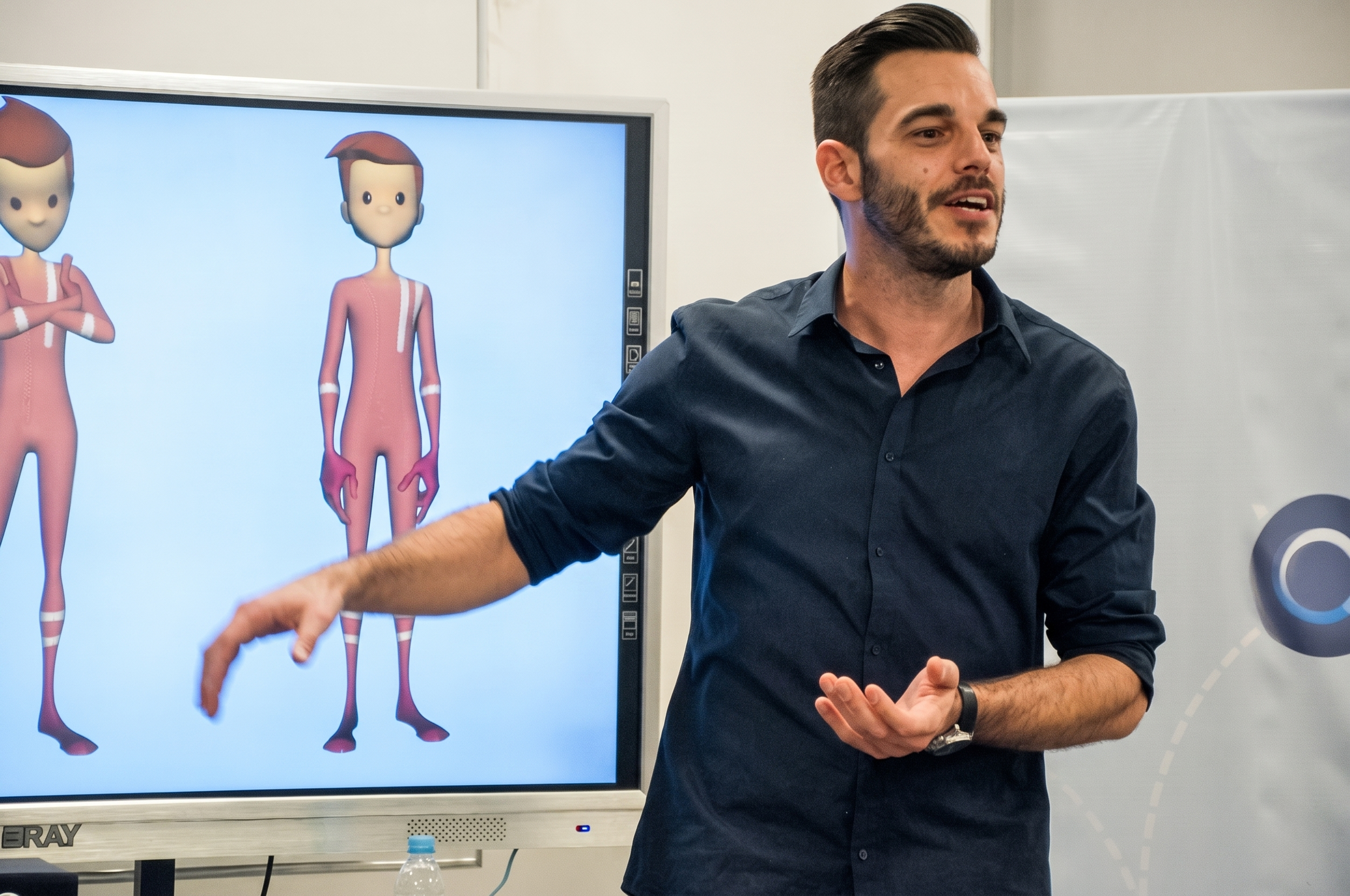 Un hombre con barba y camisa azul oscuro gesticula mientras explica frente a una pantalla que muestra figuras humanas de dibujos animados en colores rosados, en un entorno de aula o conferencia.