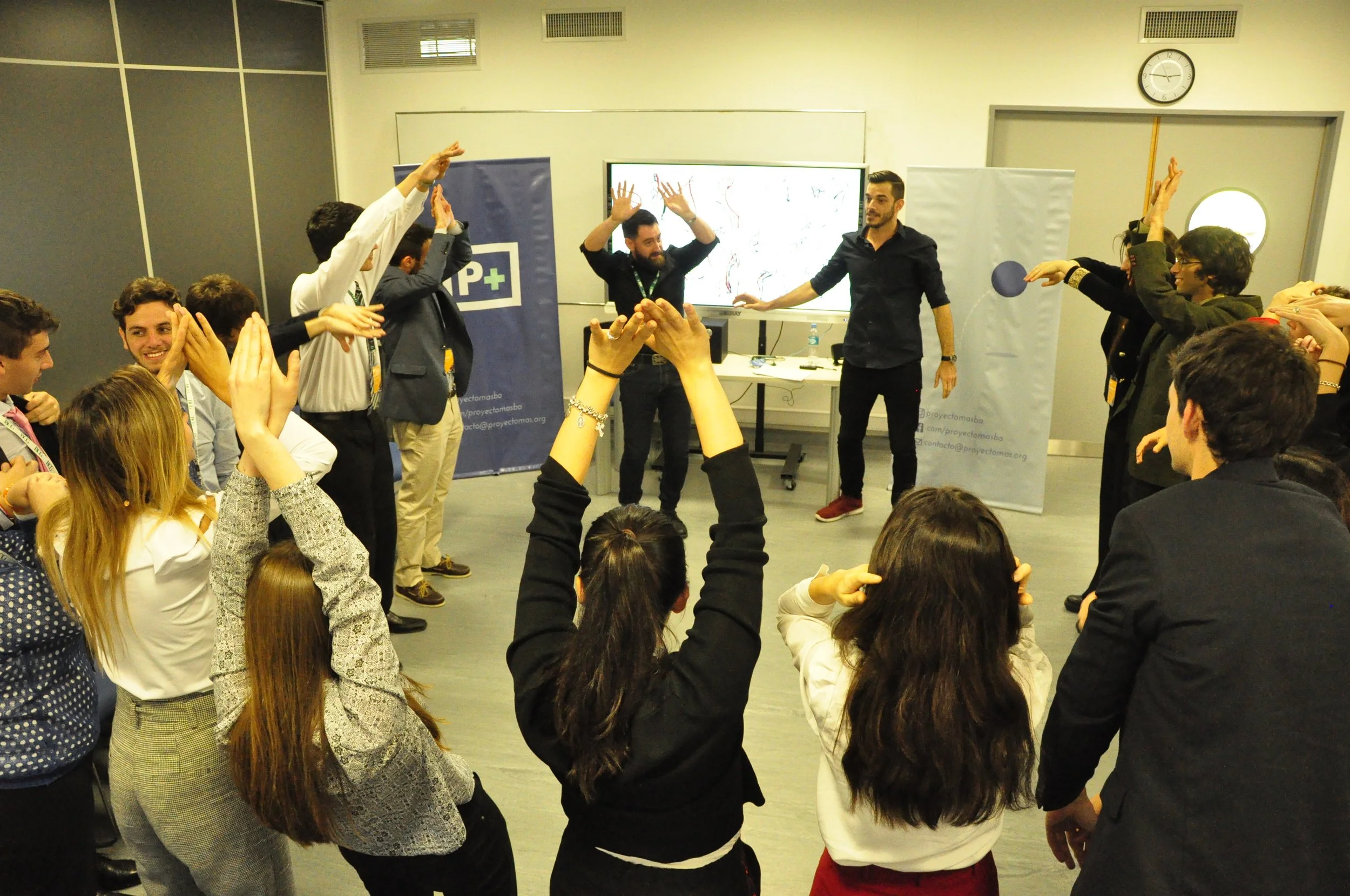 Grupo de personas participando en actividad de brinco en una sala de reuniones con pantalla, cartel y reloj.