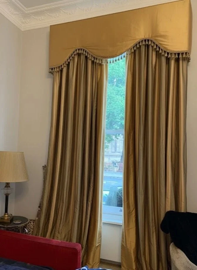 Gold-colored curtains with valance and tassel trim, partially open to reveal a window with greenery outside in a well-decorated room.