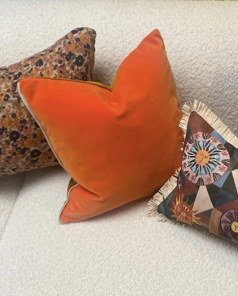 Three decorative pillows on a white textured sofa, including a floral-patterned pillow, an orange velvet pillow, and a colorful geometric and floral patterned pillow with fringe.
